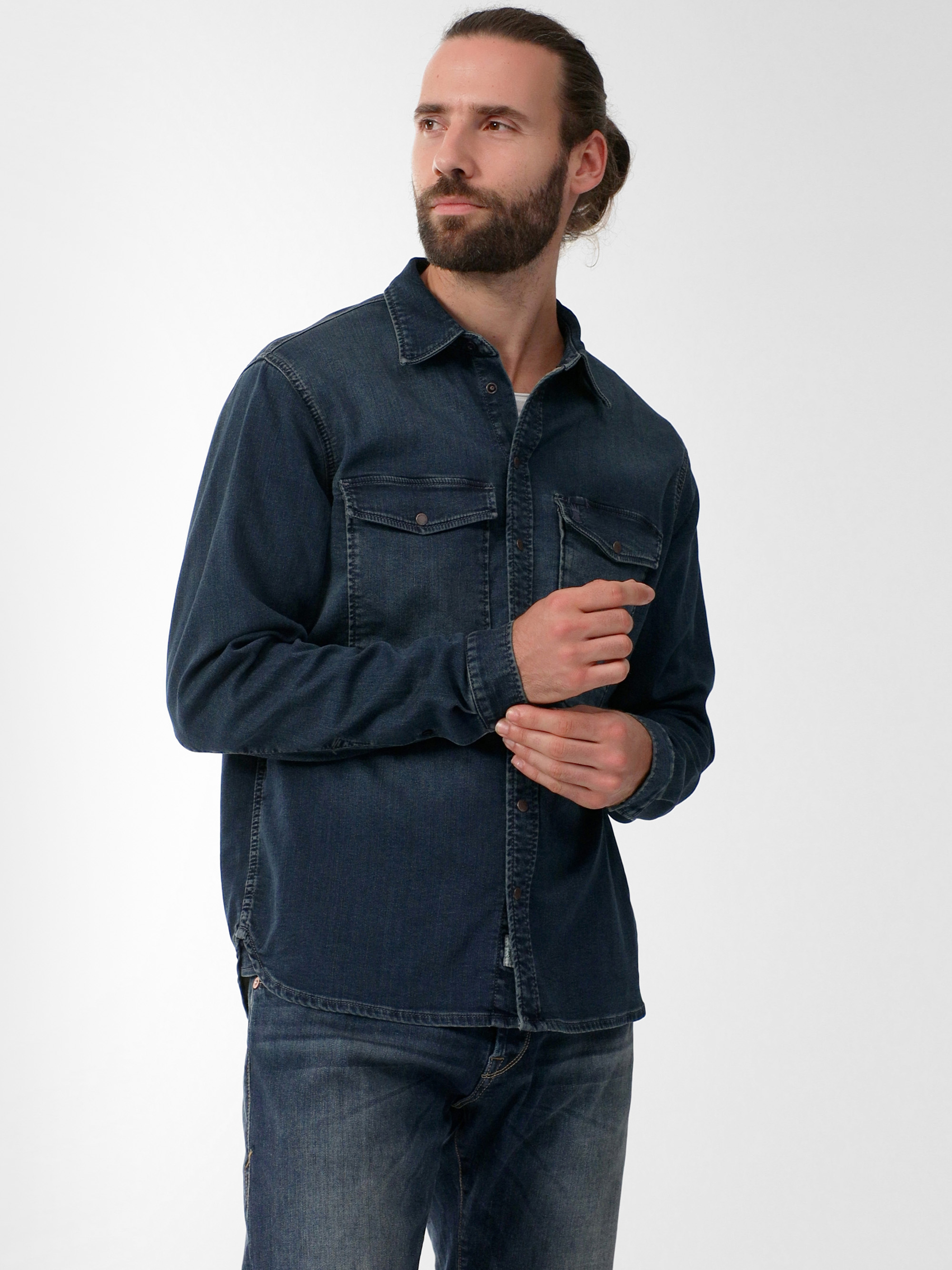 Ein Mann mit Bart und langem, zurückgebundenem Haar trägt ein dunkles Jeanshemd und Jeans. Er steht vor einem schlichten weißen Hintergrund und blickt leicht nach links, wobei eine Hand sein Hemd berührt.