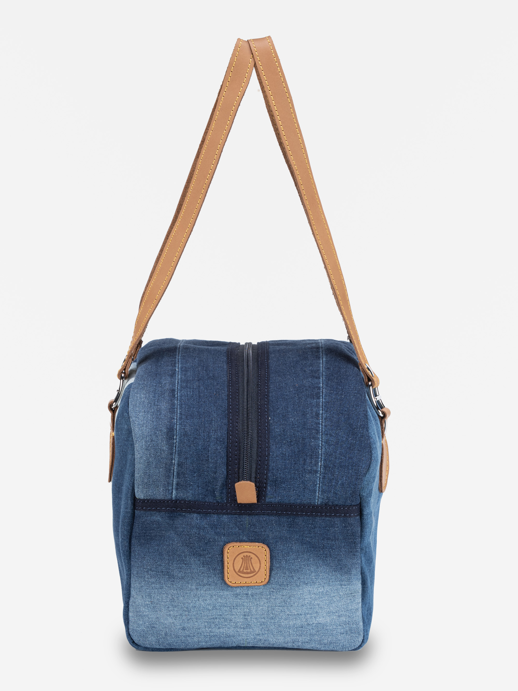 Eine Umhängetasche aus blauem Denim mit einem braunen Lederriemen, einer Reißverschlusstasche auf der Vorderseite und einem Lederaufnäher auf der Vorderseite, abgebildet auf einem schlichten weißen Hintergrund.