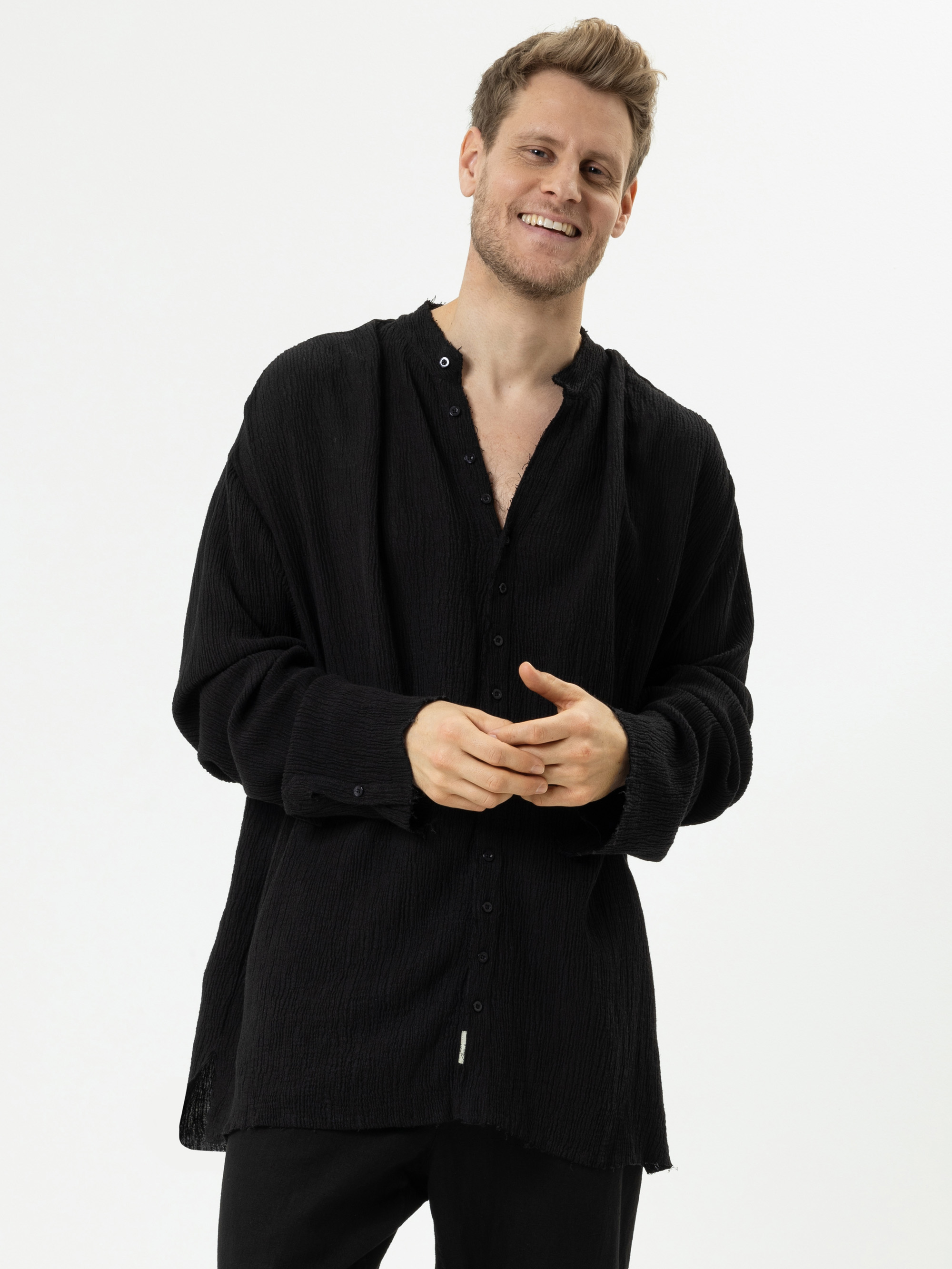 Ein lächelnder Mann mit kurzen, hellbraunen Haaren trägt ein locker sitzendes, strukturiertes schwarzes Button-up-Hemd und eine schwarze Hose und steht vor einem schlichten weißen Hintergrund.