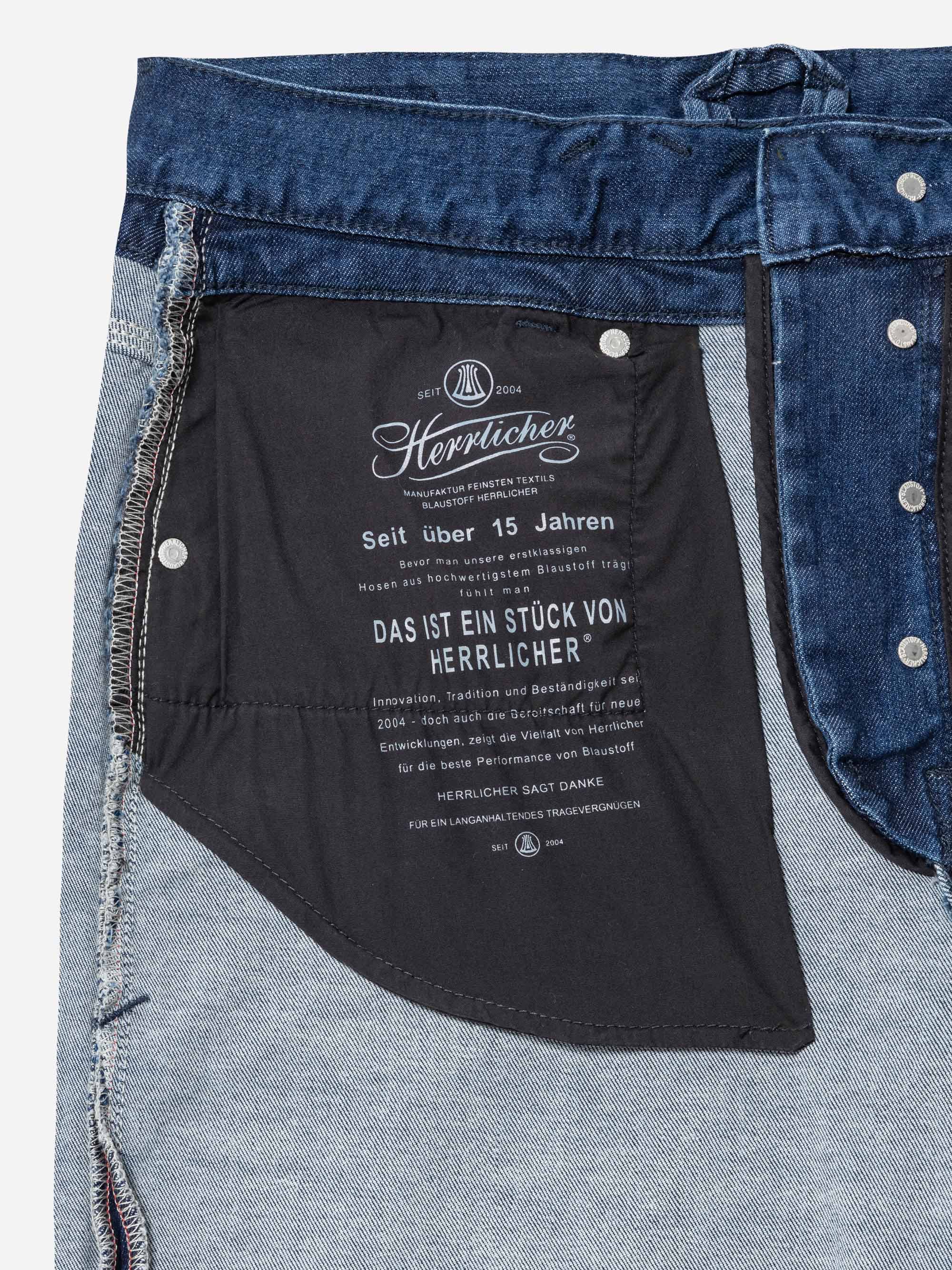 Nahaufnahme der Innenseite einer Jeans, die ein schwarzes Taschenfutter mit weiß gedrucktem deutschen Text und Branding-Details für Herrlicher zeigt, einschließlich des Firmenlogos und Informationen über das Kleidungsstück.