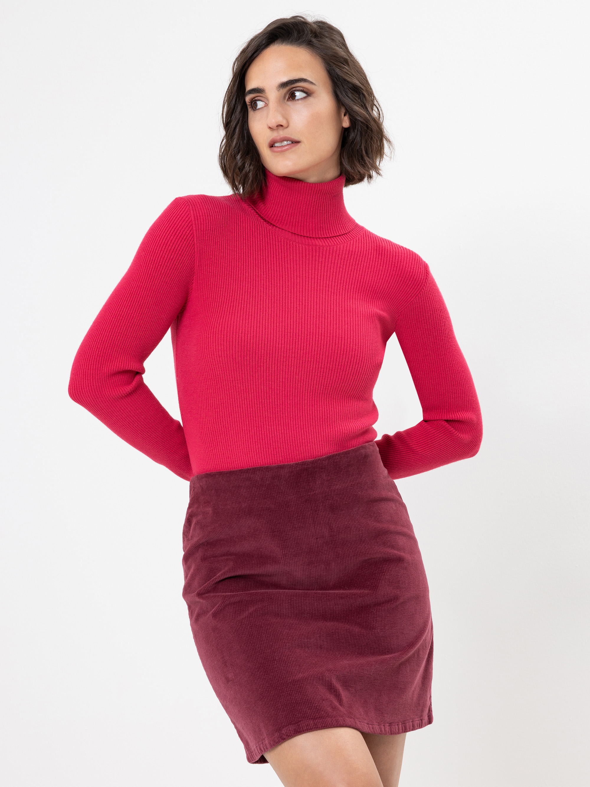 Eine Frau mit kurzen braunen Haaren trägt einen leuchtend rosafarbenen Rollkragenpullover und einen weinroten Rock mit hoher Taille. Sie steht vor einem schlichten weißen Hintergrund und schaut nach links.
