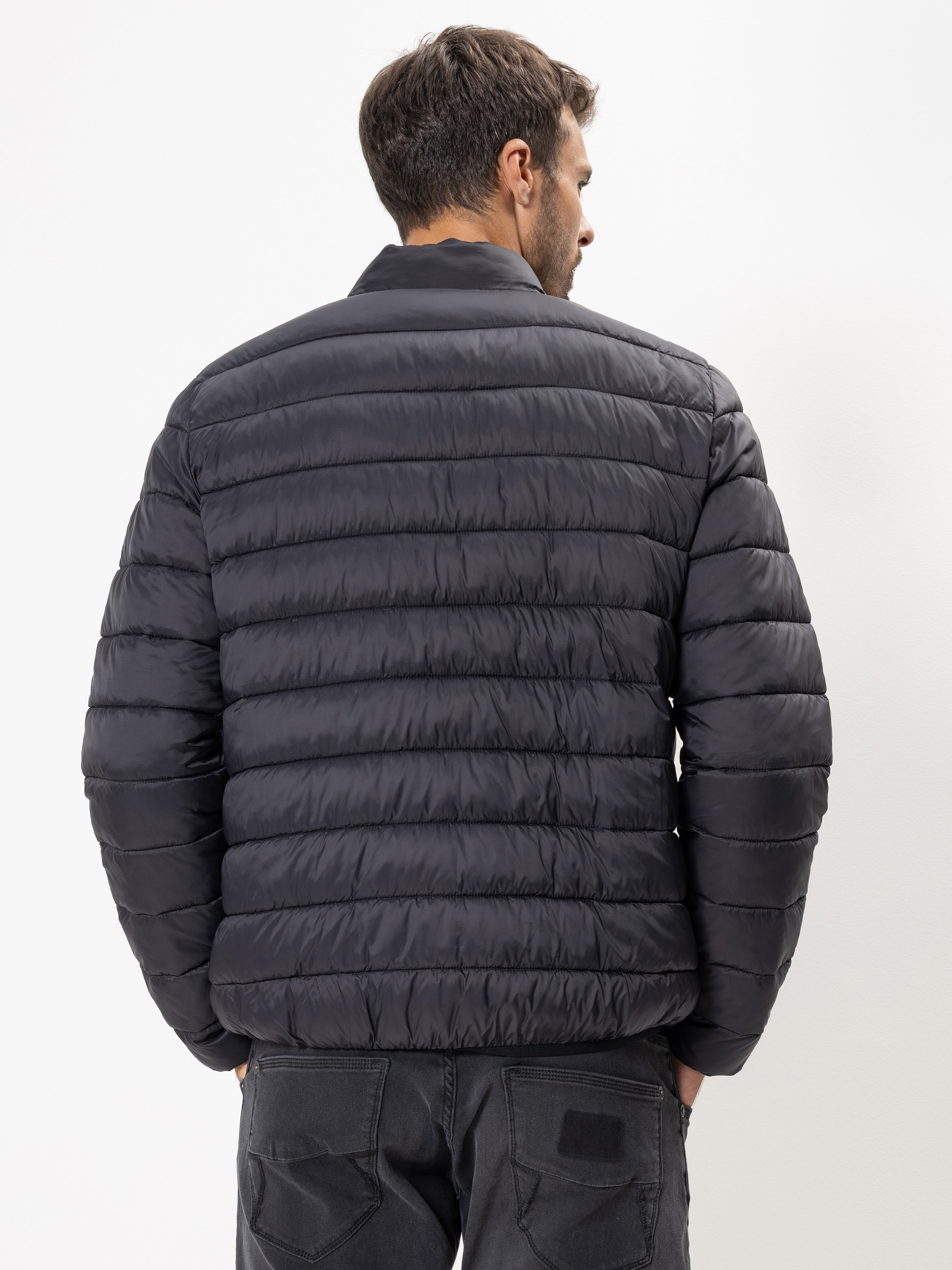 Ein Mann mit kurzen braunen Haaren wird von hinten gezeigt. Er trägt eine schwarze Steppjacke und dunkle Jeans und steht vor einem schlichten weißen Hintergrund.