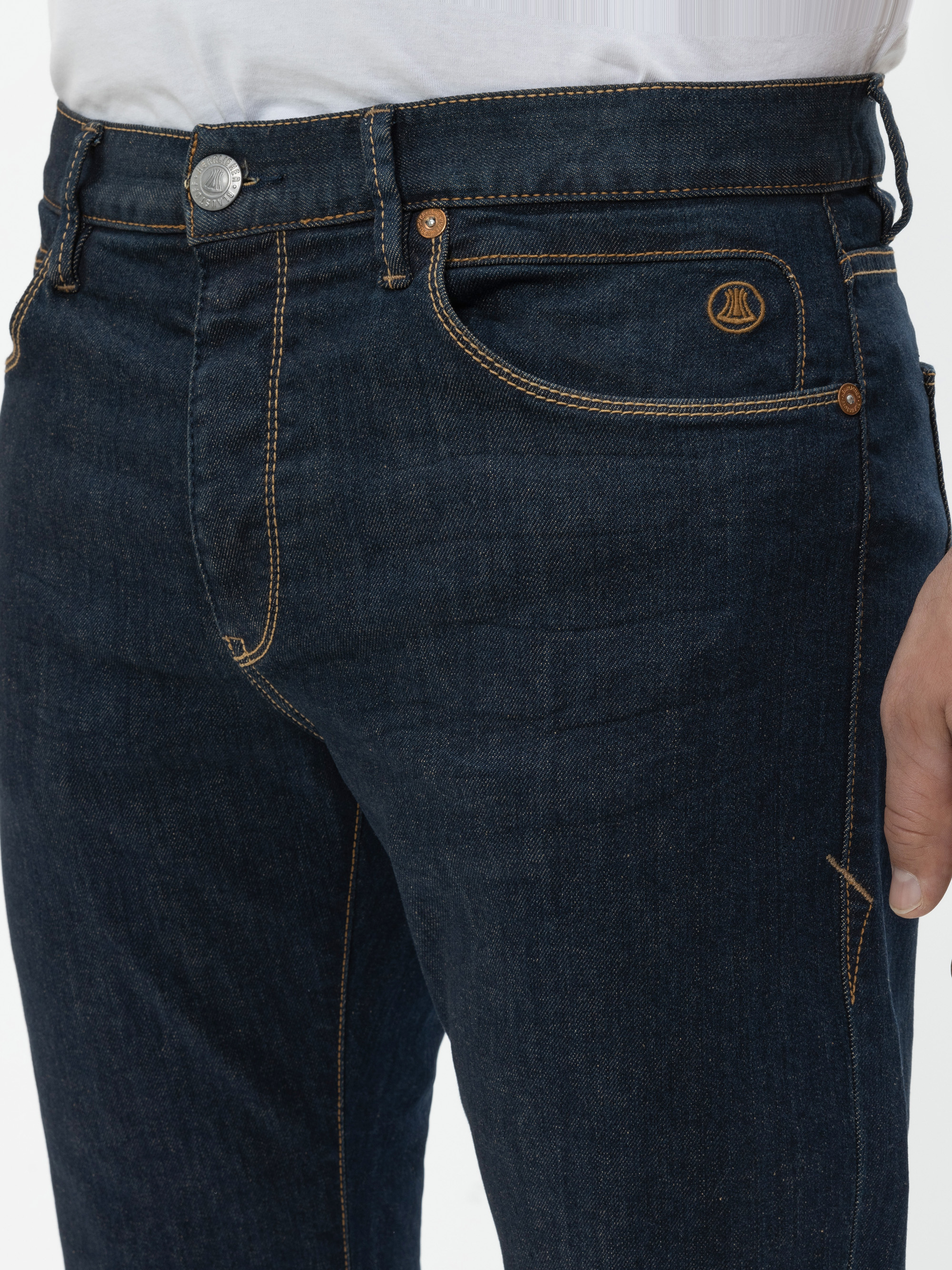 Eine Person trägt eine dunkelblaue Jeans mit gelben Nähten und ein weißes Hemd. Die Jeans hat einen Metallknopf und ein kleines gesticktes Logo über der rechten Tasche. Nur die Taille und die Oberschenkel sind sichtbar.