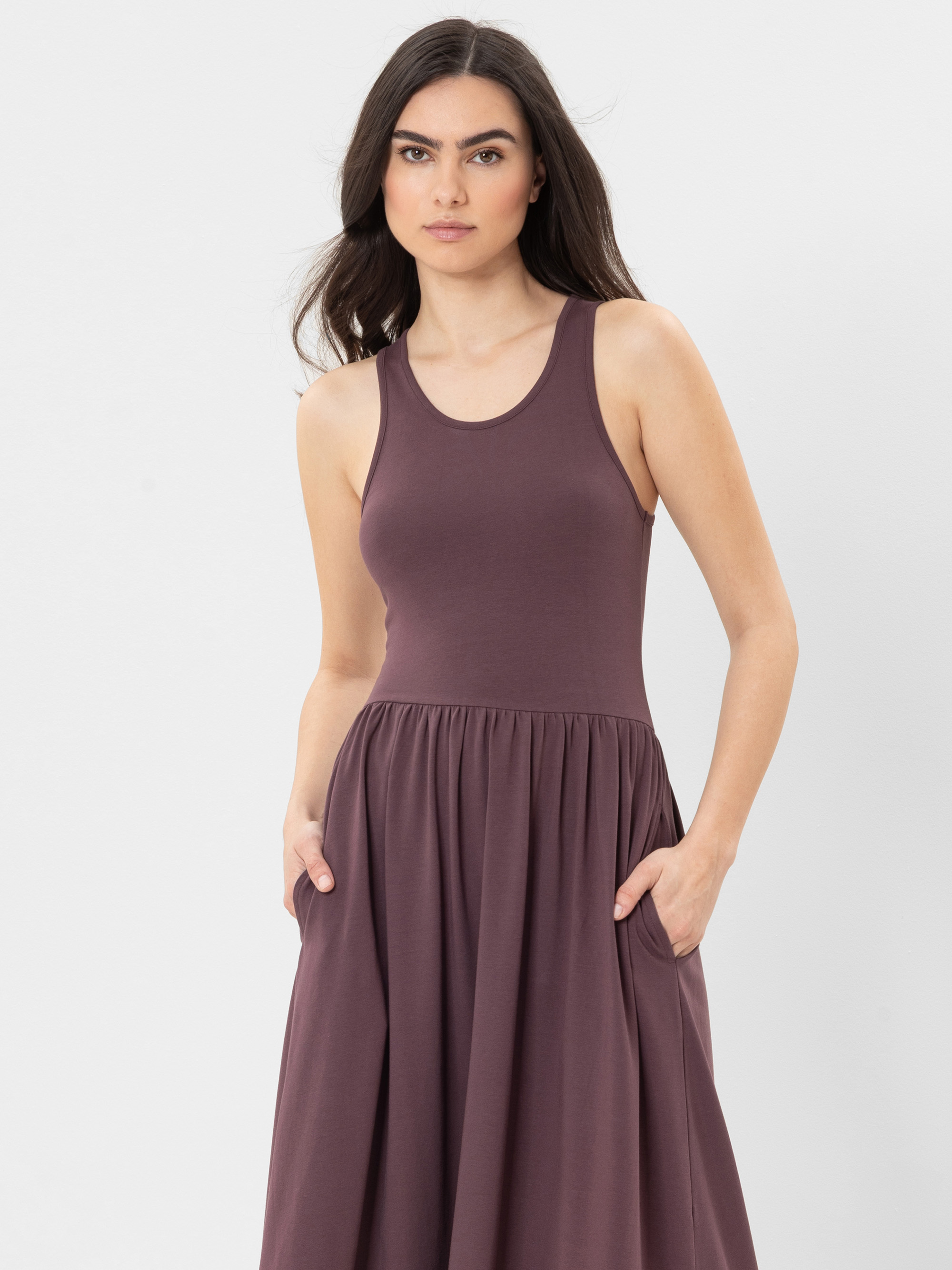 Eine Frau mit langen dunklen Haaren trägt ein ärmelloses, lilafarbenes, tailliertes Kleid mit Taschen und steht vor einem schlichten weißen Hintergrund.