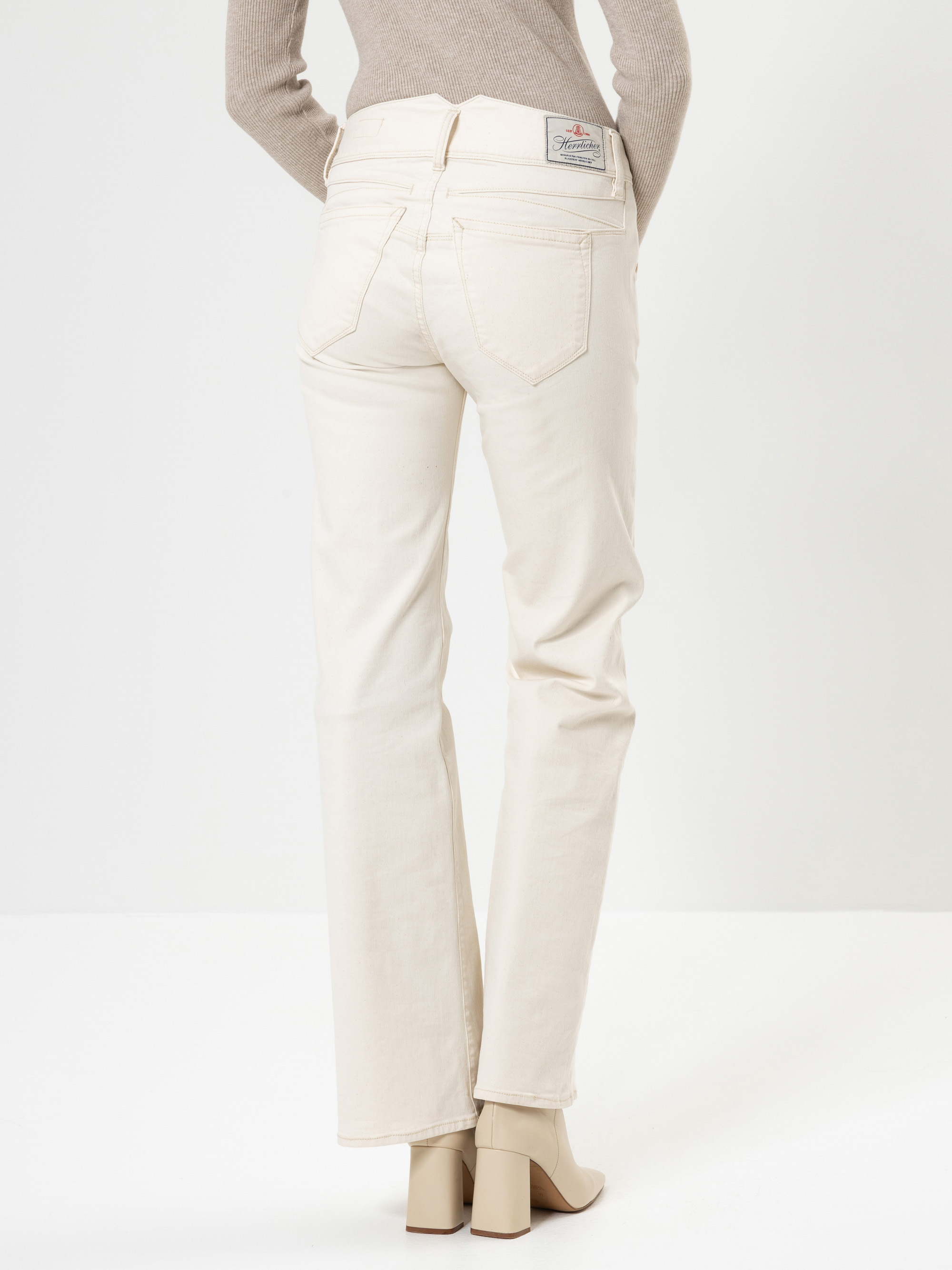 Eine Person, die cremefarbene Jeans, einen beigen gerippten Pullover und beigefarbene hochhackige Stiefel trägt, steht mit dem Rücken zur Kamera vor einem schlichten weißen Hintergrund.