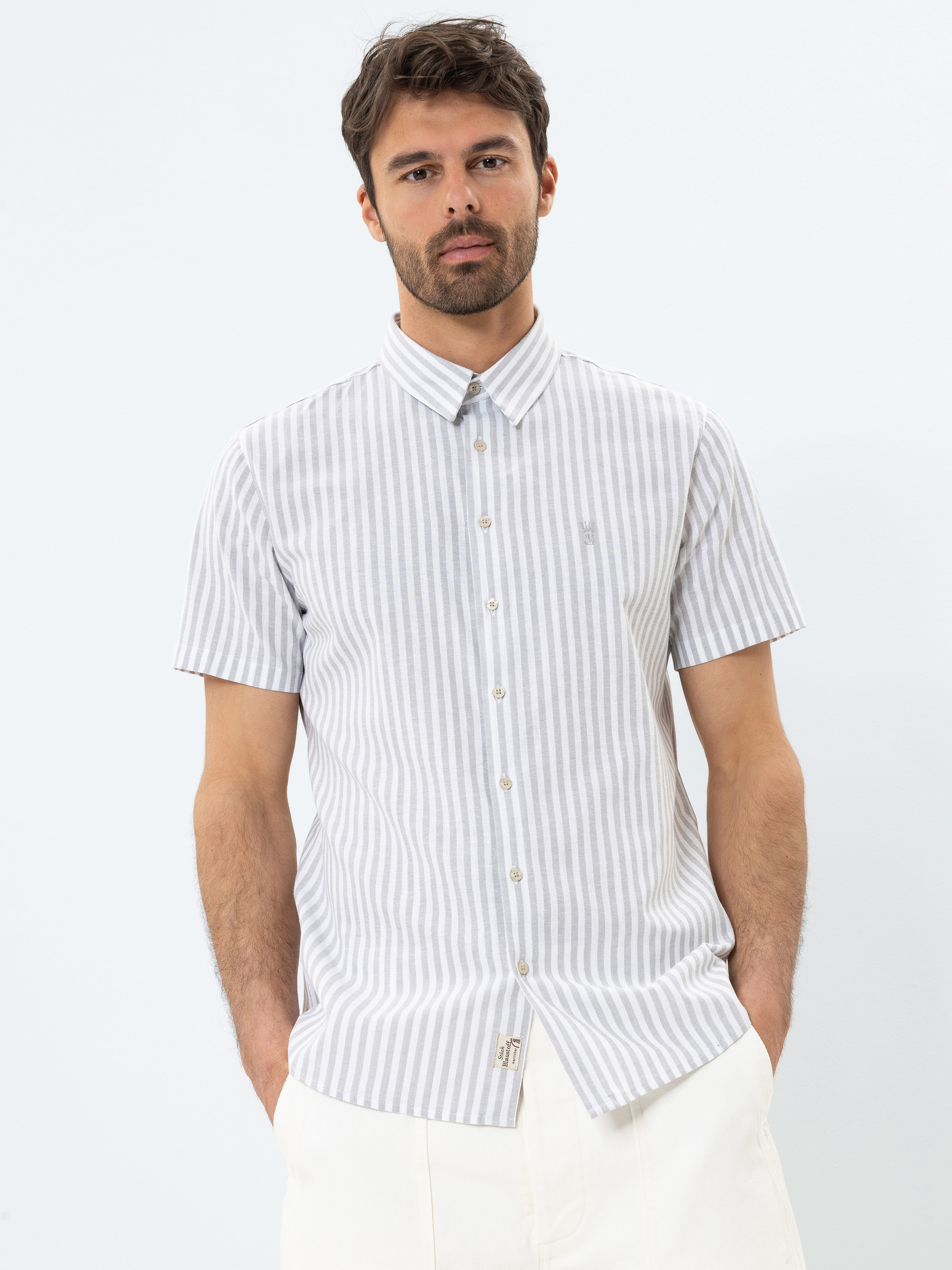 Ein Mann mit kurzen braunen Haaren und Bart trägt ein kurzärmeliges, hellgrau-weiß vertikal gestreiftes Button-up-Hemd und eine weiße Hose. Er steht vor einem schlichten weißen Hintergrund und hat die Hände in den Taschen.