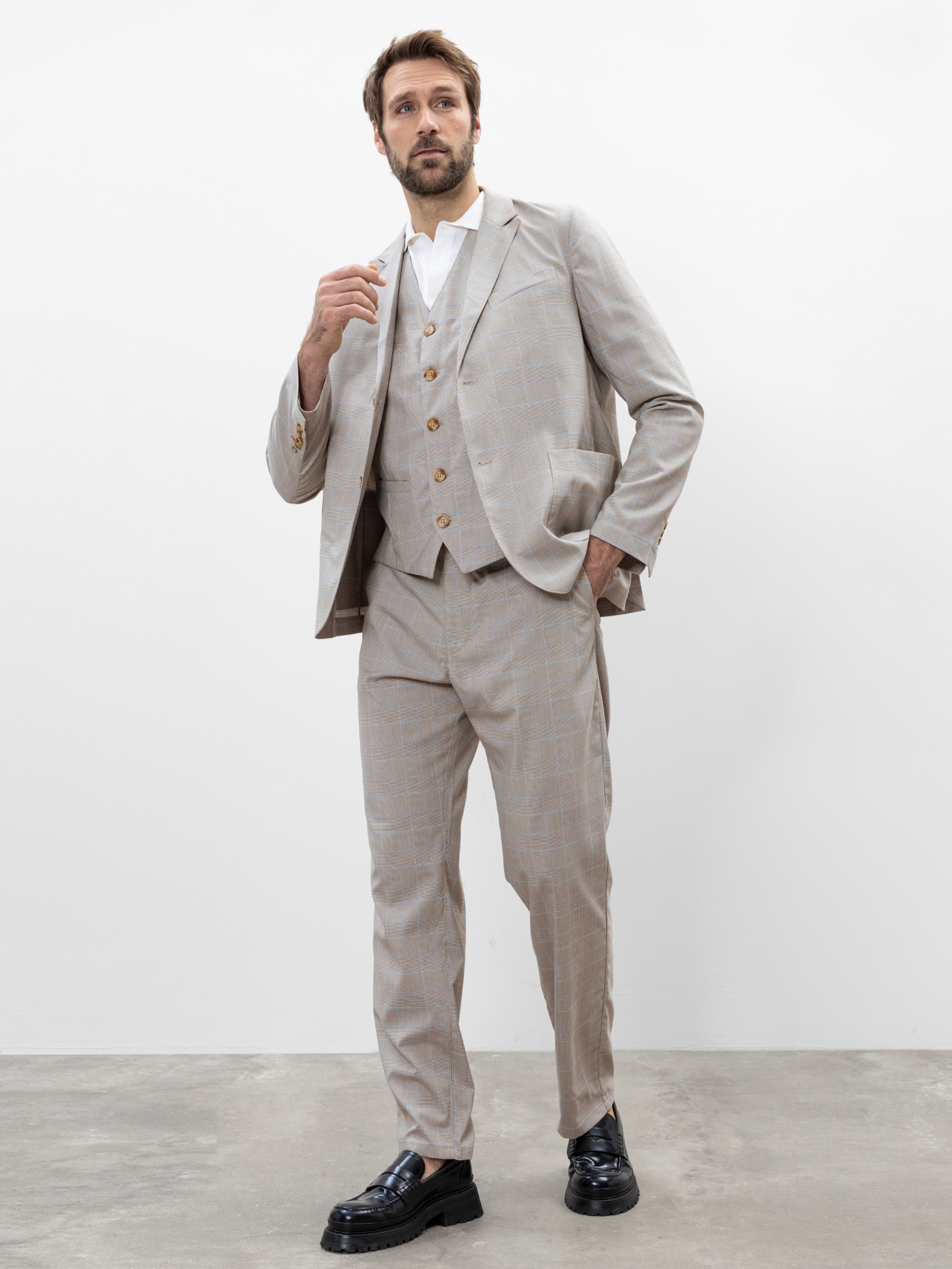 Ein Mann steht auf einem Betonboden vor einer schlichten weißen Wand. Er trägt einen hellbeigen karierten Anzug mit passender Weste, ein weißes Hemd und schwarze Anzugschuhe, eine Hand am Kragen, die andere in der Tasche.