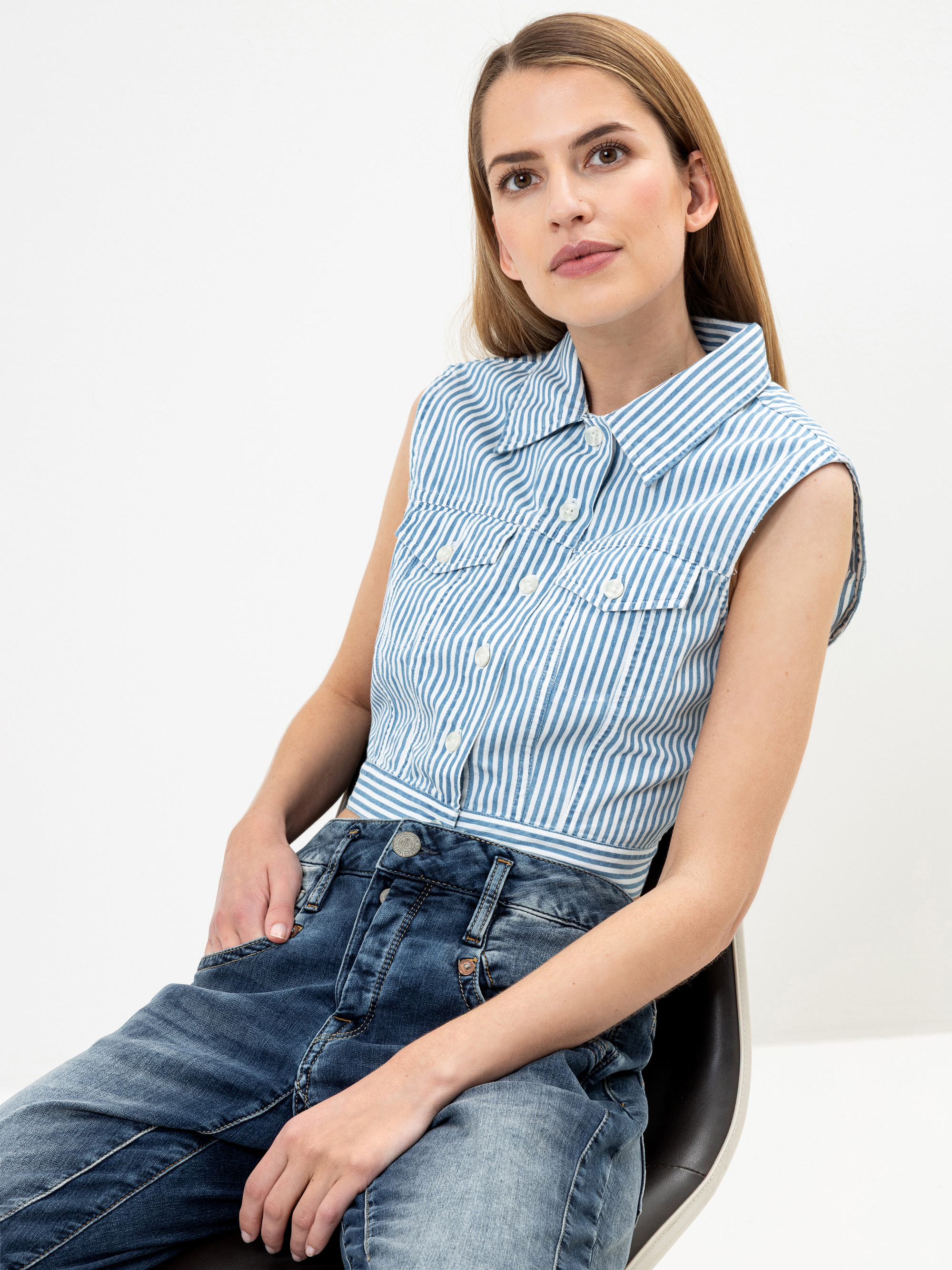 Eine Frau mit langem hellbraunem Haar sitzt auf einem Stuhl, trägt ein ärmelloses blau-weiß gestreiftes Button-up-Hemd und hochgekrempelte Bluejeans und blickt mit neutraler Miene leicht nach oben.