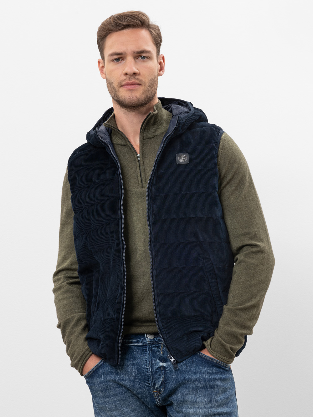 Ein Mann mit kurzen braunen Haaren trägt eine dunkelblaue Steppweste über einem olivgrünen Pullover mit Reißverschluss und blaue Jeans. Er steht vor einem schlichten weißen Hintergrund und hat die Hände in den Taschen.