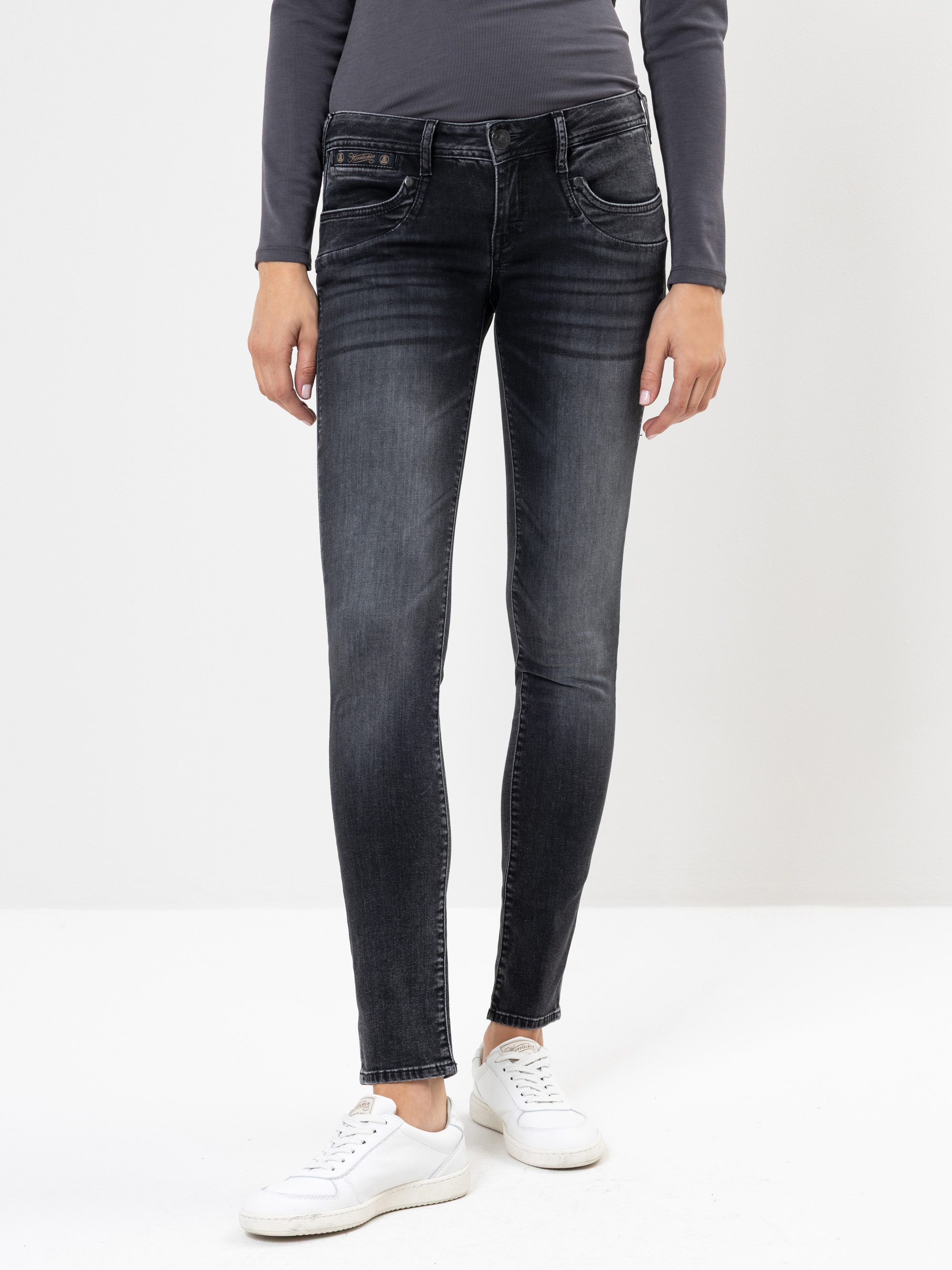 Eine Person, die dunkel gewaschene Röhrenjeans, ein langärmeliges dunkelgraues Oberteil und weiße Turnschuhe trägt und vor einem schlichten weißen Hintergrund steht. Das Bild ist an den Schultern abgeschnitten.