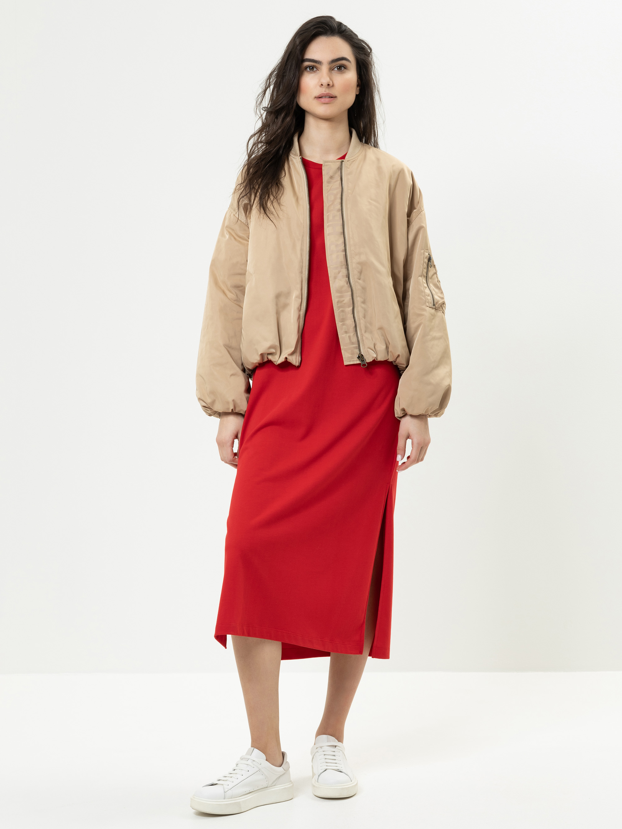 Eine Frau steht vor einem weißen Hintergrund und trägt eine hellbraune Bomberjacke über einem roten Midikleid mit Seitenschlitz, kombiniert mit weißen Turnschuhen.