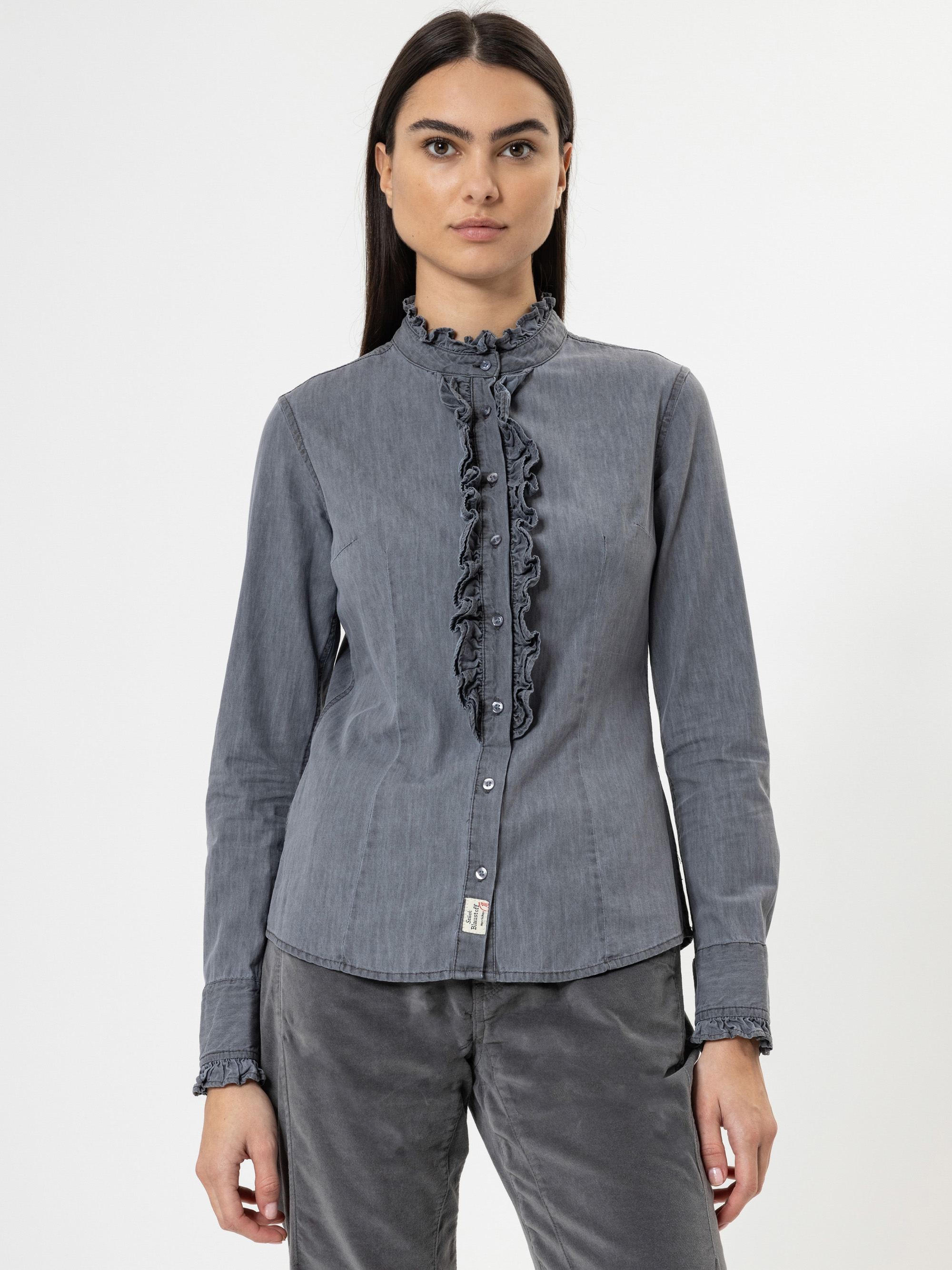 Eine Frau mit langen dunklen Haaren trägt eine graue, langärmelige Bluse mit Rüschen an der Knopfleiste und den Manschetten, gepaart mit einer grauen Hose, vor einem schlichten weißen Hintergrund.