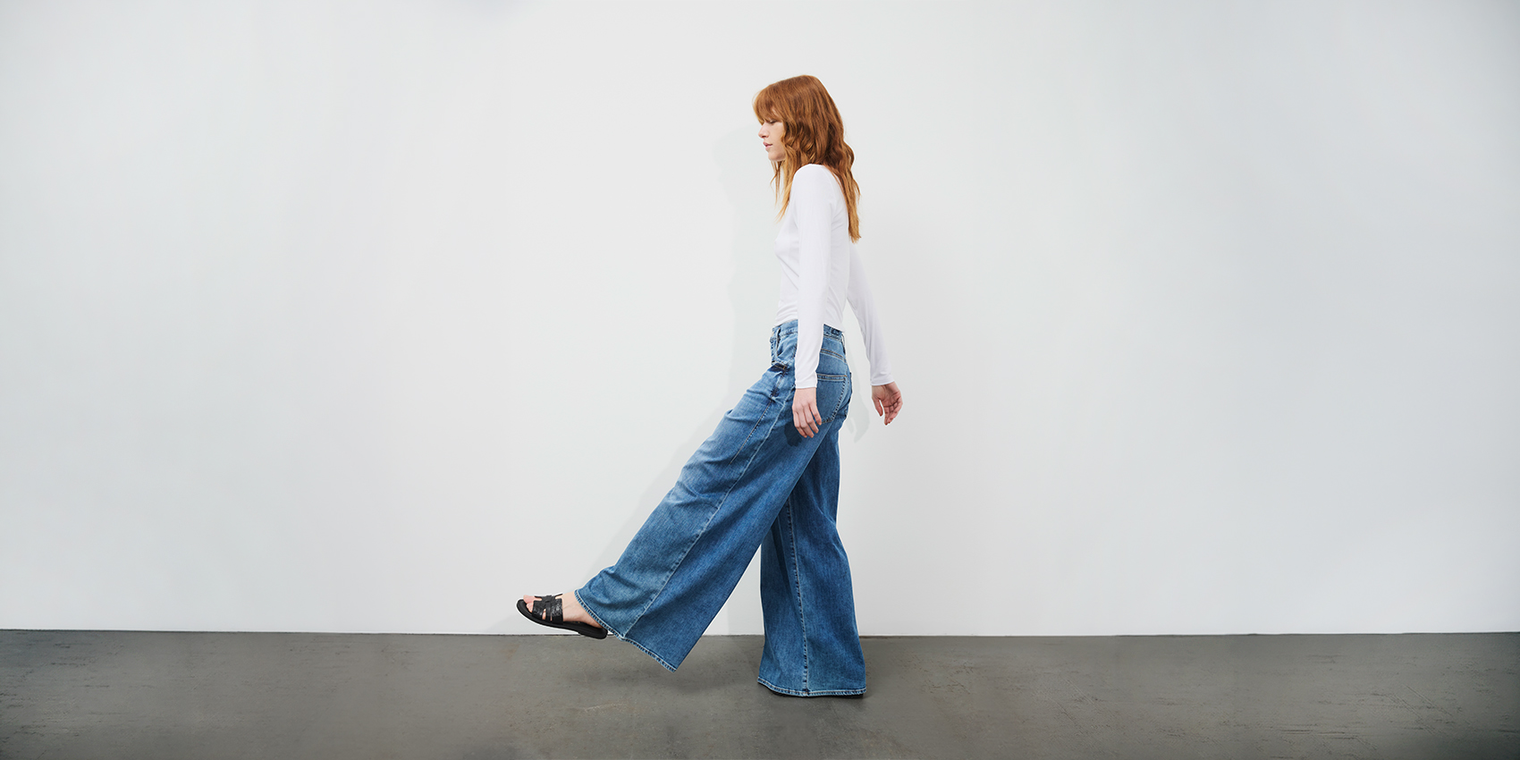 Eine Frau mit langen roten Haaren, die ein weißes Langarmshirt und eine blaue Jeans mit weitem Bein trägt, geht seitwärts vor einem schlichten weißen Hintergrund auf einem dunklen Boden.