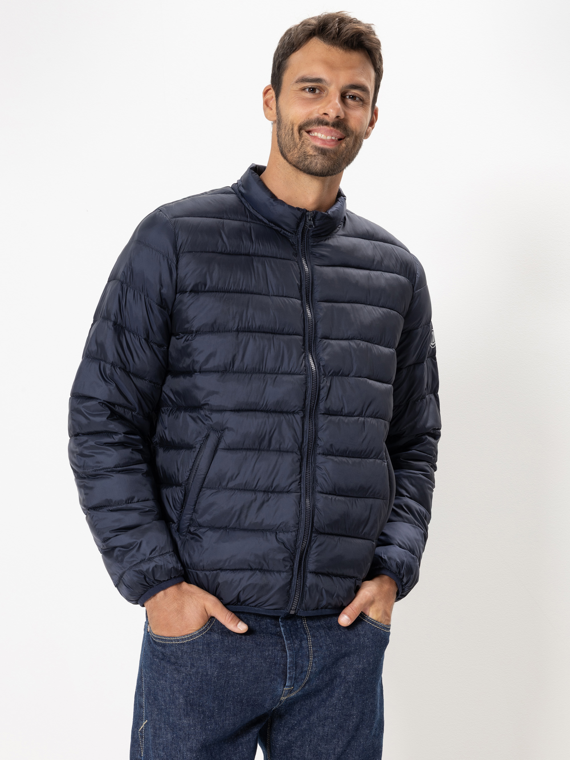 Ein Mann, der eine dunkelblaue Pufferjacke und blaue Jeans trägt, steht vor einem schlichten weißen Hintergrund und lächelt mit den Händen in seinen Taschen.