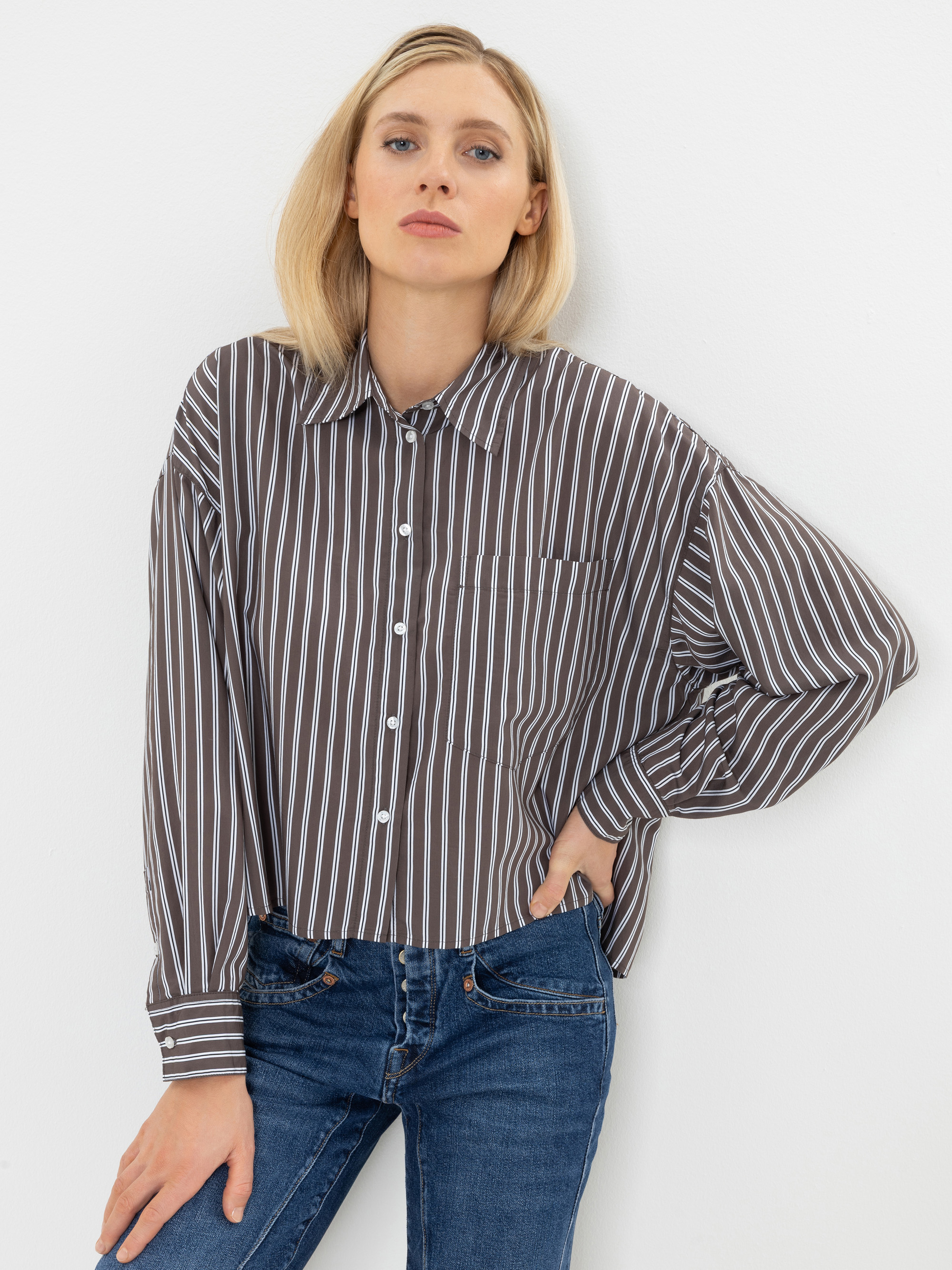 Eine Person mit blonden Haaren trägt ein braun-weiß gestreiftes Hemd mit Knöpfen und eine blaue Jeans. Sie steht vor einer weißen Wand, eine Hand in der Hosentasche, die andere an der Seite.