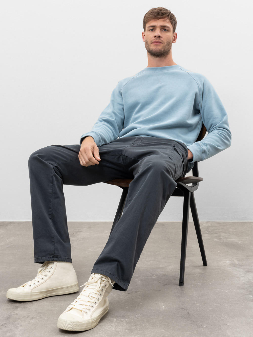 Ein Mann in einem hellblauen Sweatshirt, einer dunkelgrauen Hose und weißen Turnschuhen mit hohen Absätzen sitzt lässig auf einem Stuhl vor einem schlichten, hellen Hintergrund.