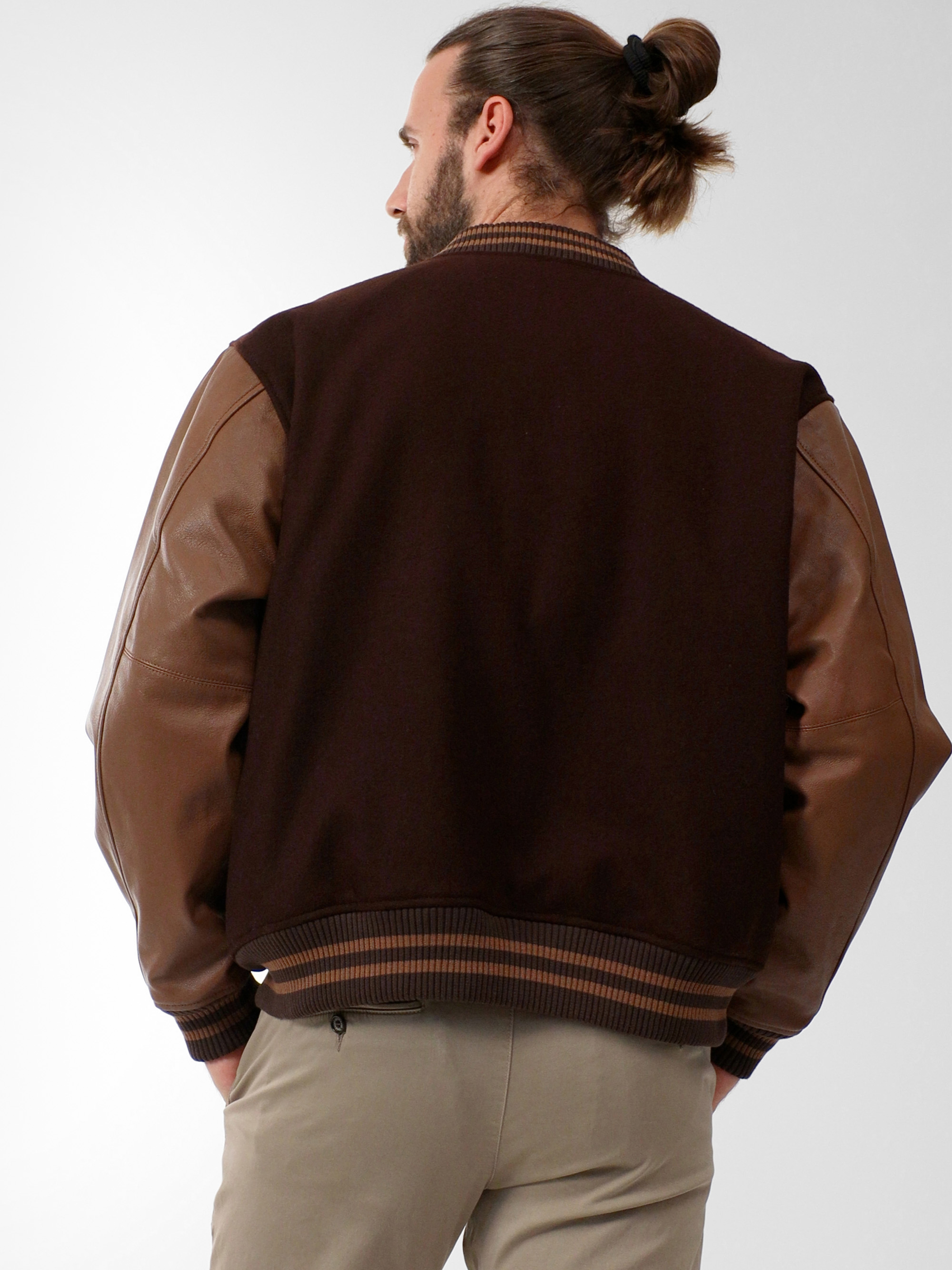 Man sieht einen Mann mit einem braunen Dutt von hinten, der eine dunkelbraune Jacke mit hellbraunen Lederärmeln und eine hellbeige Hose trägt. Der Hintergrund ist einfarbig und hell.