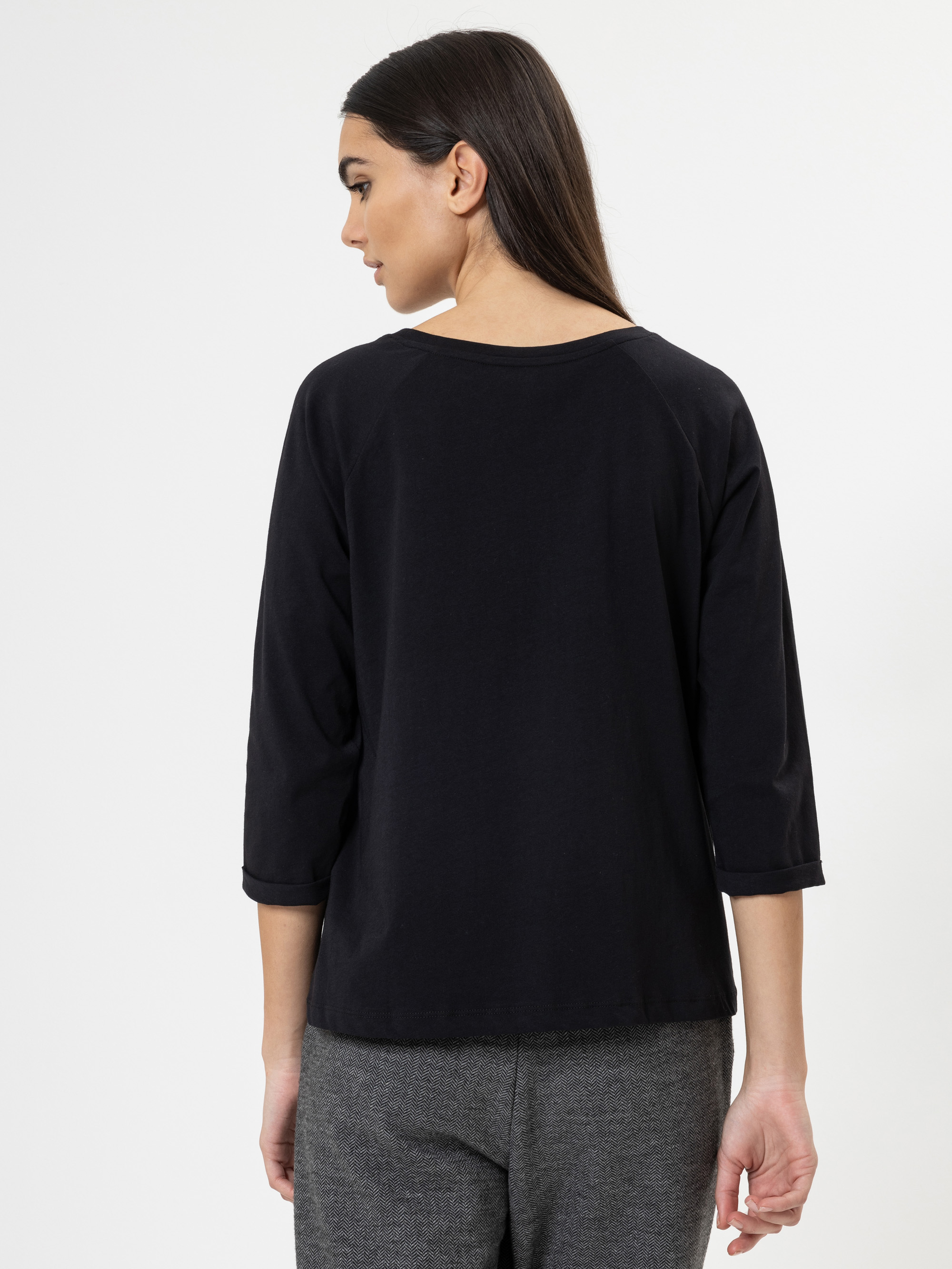 Frau mit langen braunen Haaren, die ein schwarzes dreiviertelärmeliges Oberteil und eine graue Hose trägt und mit dem Rücken zur Kamera vor einem schlichten weißen Hintergrund steht.