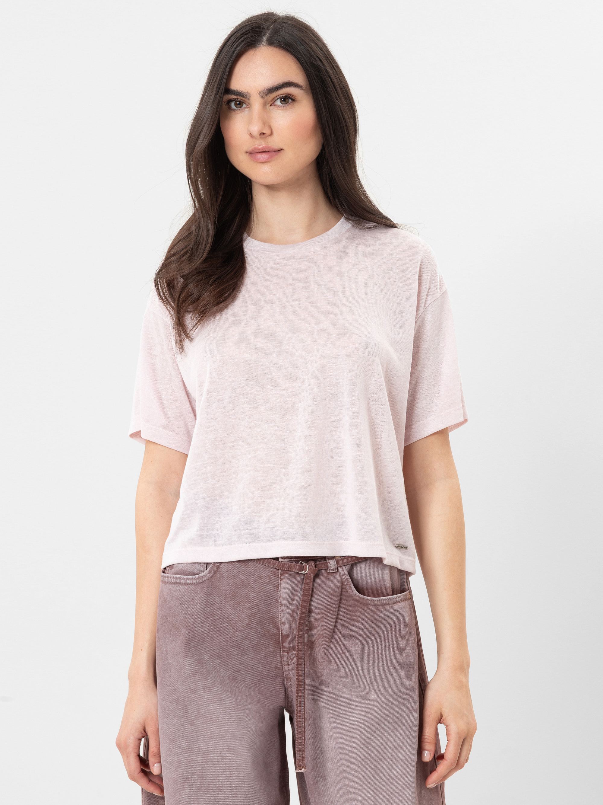 Eine Frau mit langen braunen Haaren trägt ein lockeres, hellrosa T-Shirt mit kurzen Ärmeln und eine hochgekrempelte lilafarbene Jeans und steht vor einem schlichten weißen Hintergrund.