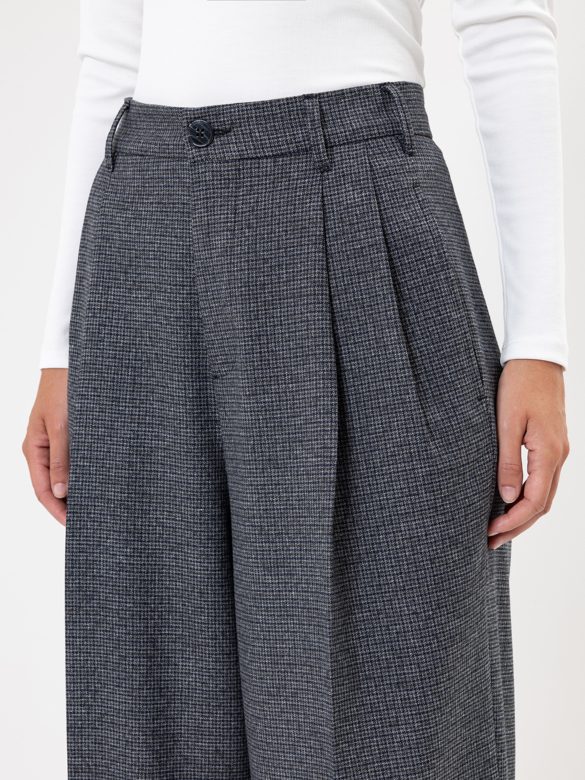 Eine Person trägt eine graue, karierte Hose mit hoher Taille und weitem Bein, die in Falten gelegt ist, und ein weißes, langärmeliges Oberteil. Das Bild konzentriert sich auf die Taille und die Oberschenkel und zeigt die Textur und die Passform der Hos