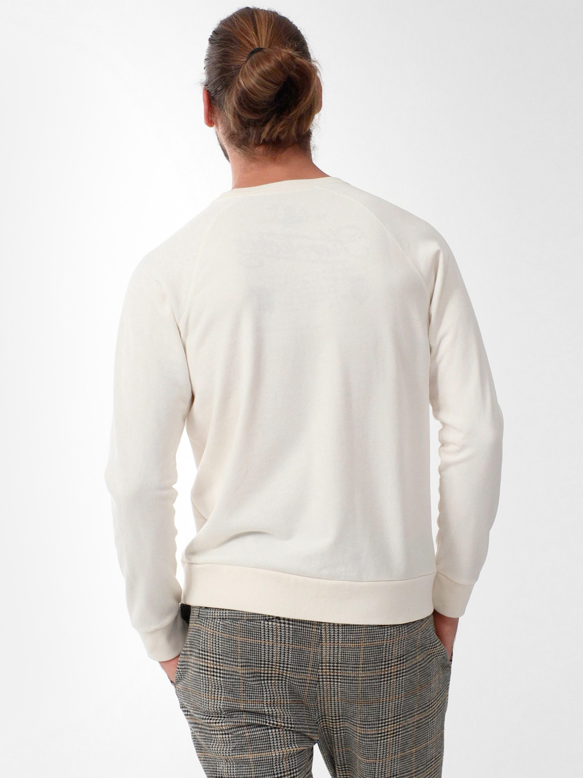 Ein Mann mit einer Duttfrisur ist von hinten zu sehen. Er trägt ein cremefarbenes, langärmeliges Sweatshirt und eine grau karierte Hose vor einem schlichten weißen Hintergrund.