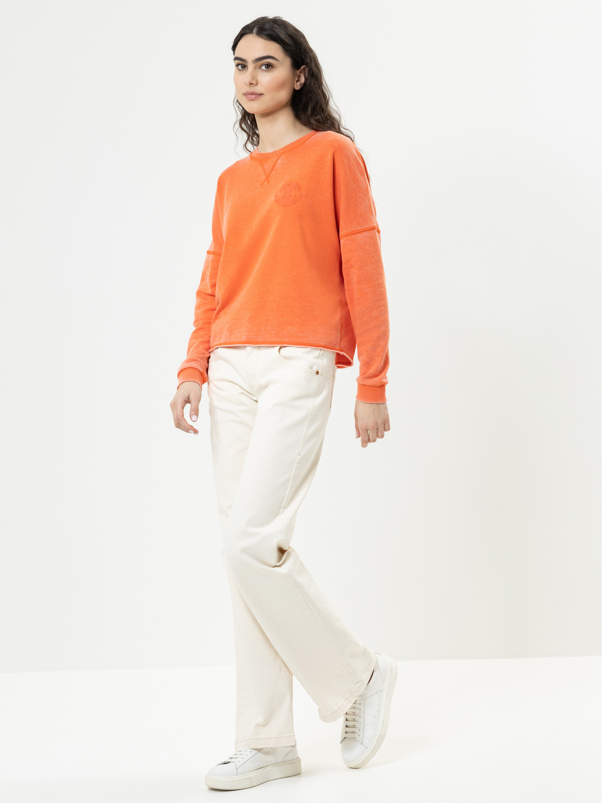 Eine Frau steht vor einem weißen Hintergrund und trägt ein orangefarbenes langärmeliges Sweatshirt, eine gebrochen weiße Hose und weiße Turnschuhe. Sie hat langes dunkles Haar und schaut mit entspanntem Gesichtsausdruck leicht zur Seite.