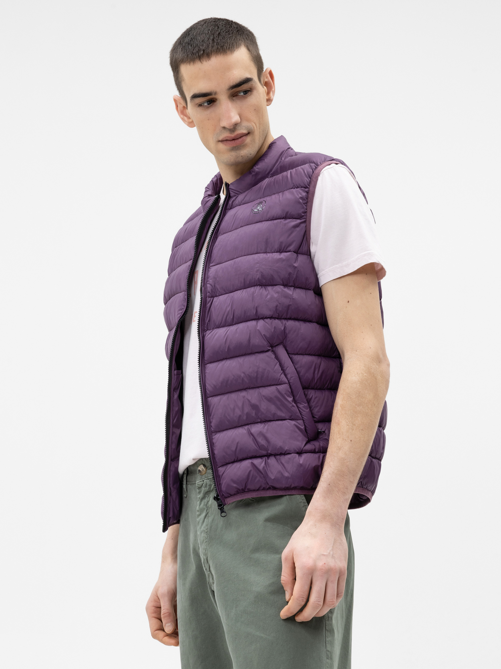 Ein Mann, der eine lilafarbene Pufferweste über einem weißen T-Shirt und eine grüne Hose trägt, steht vor einem schlichten weißen Hintergrund und schaut nach links.