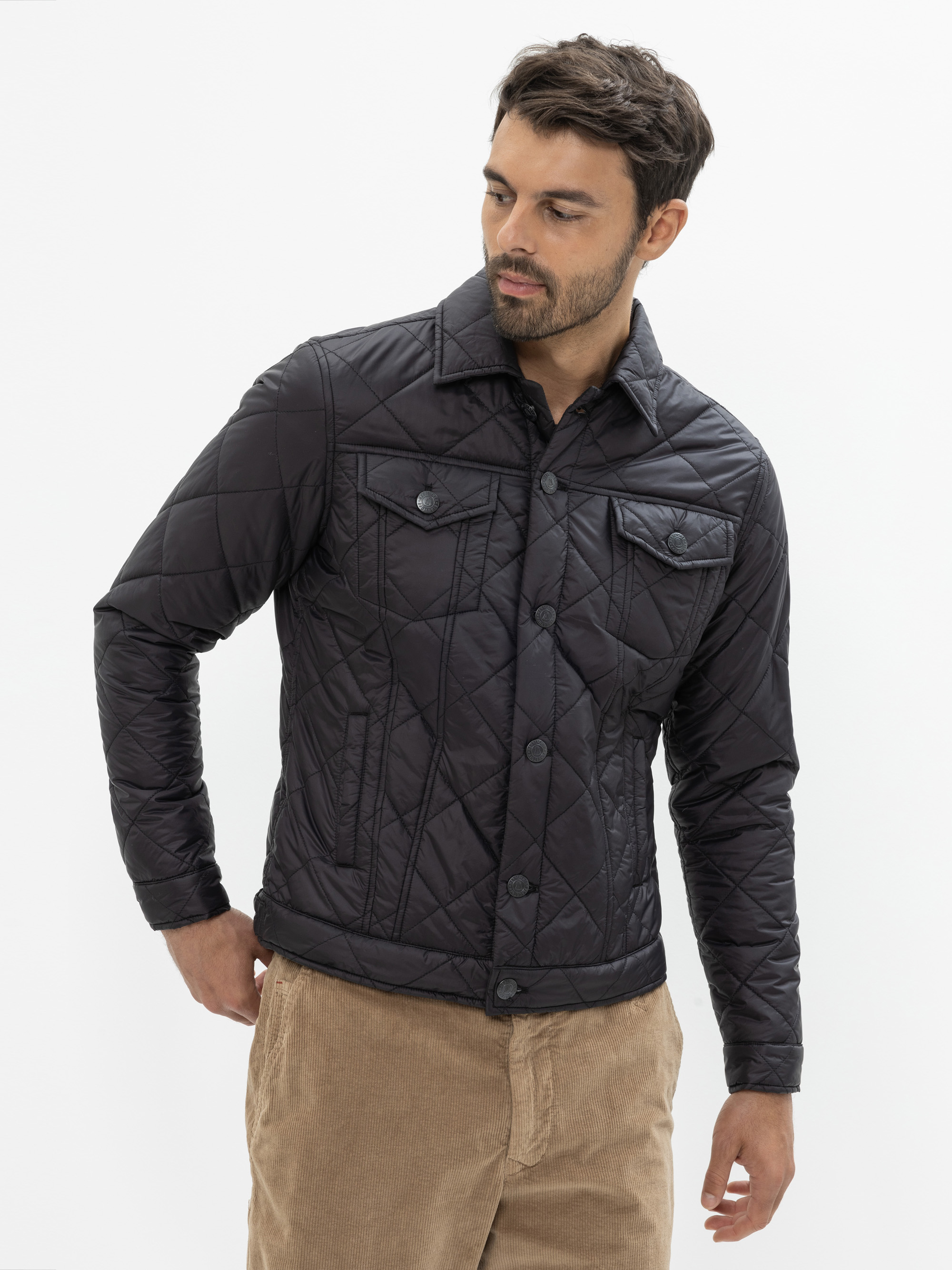 Ein Mann, der eine schwarze Steppjacke mit Druckknöpfen und Klappentaschen trägt, steht vor einem weißen Hintergrund und schaut leicht nach links. Er trägt außerdem eine hellbraune Cordhose.