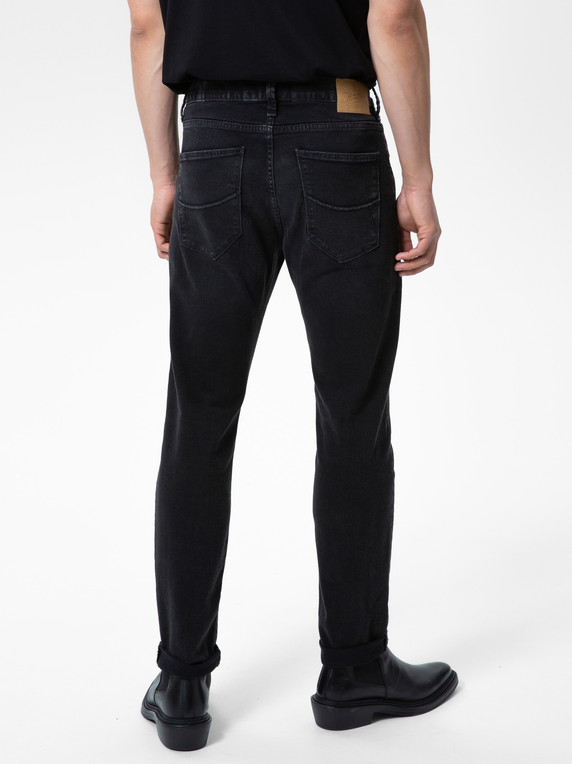 Eine Person steht mit dem Gesicht zur Wand und trägt ein schwarzes T-Shirt, schwarze Röhrenjeans mit Gesäßtaschen und schwarze Schuhe. Der Hintergrund ist einfarbig weiß.