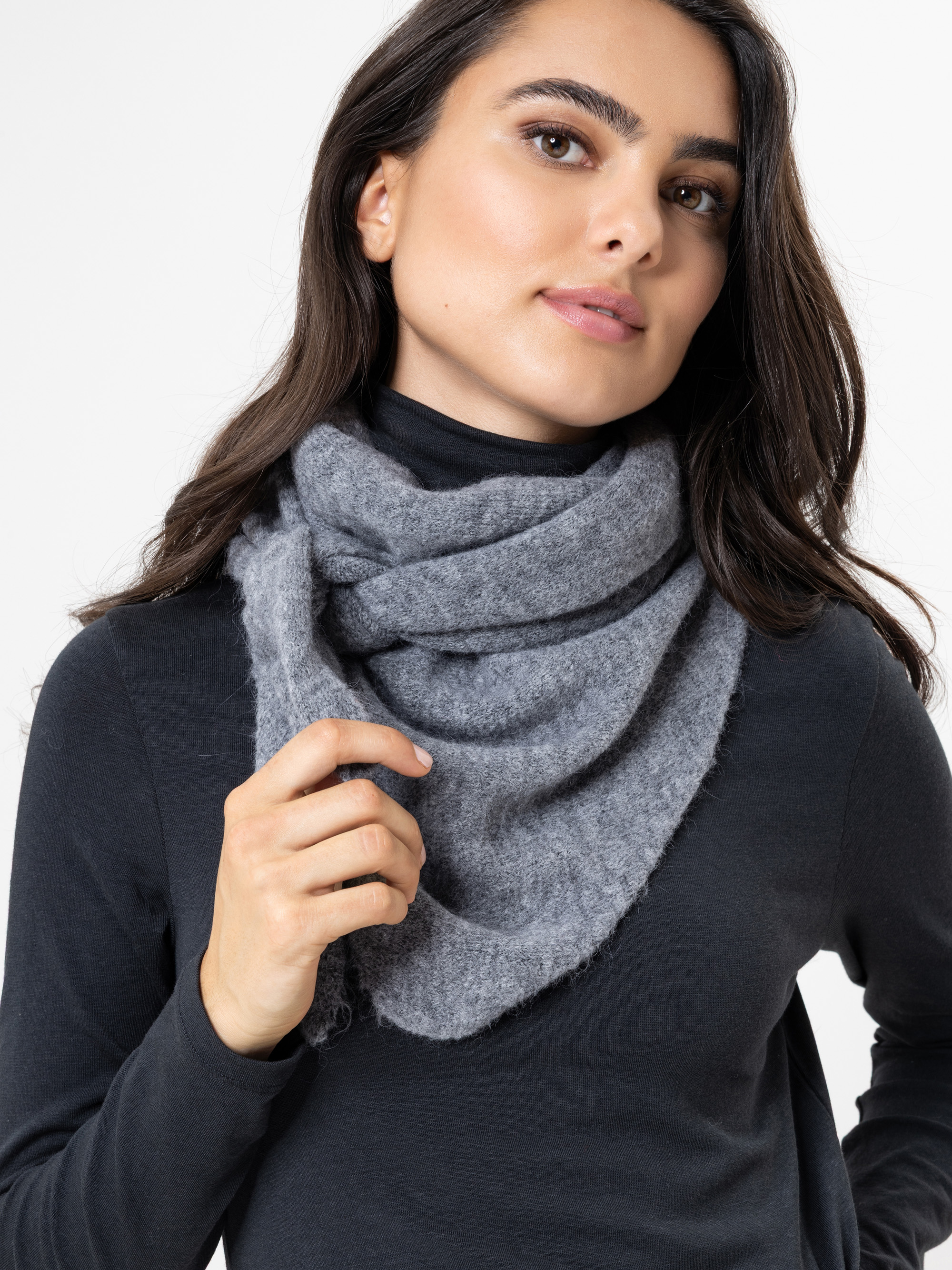 Eine Frau mit langen dunklen Haaren, die einen grauen Wollschal und ein schwarzes langärmeliges Oberteil trägt, blickt leicht zur Seite und hat einen neutralen Ausdruck vor einem weißen Hintergrund.