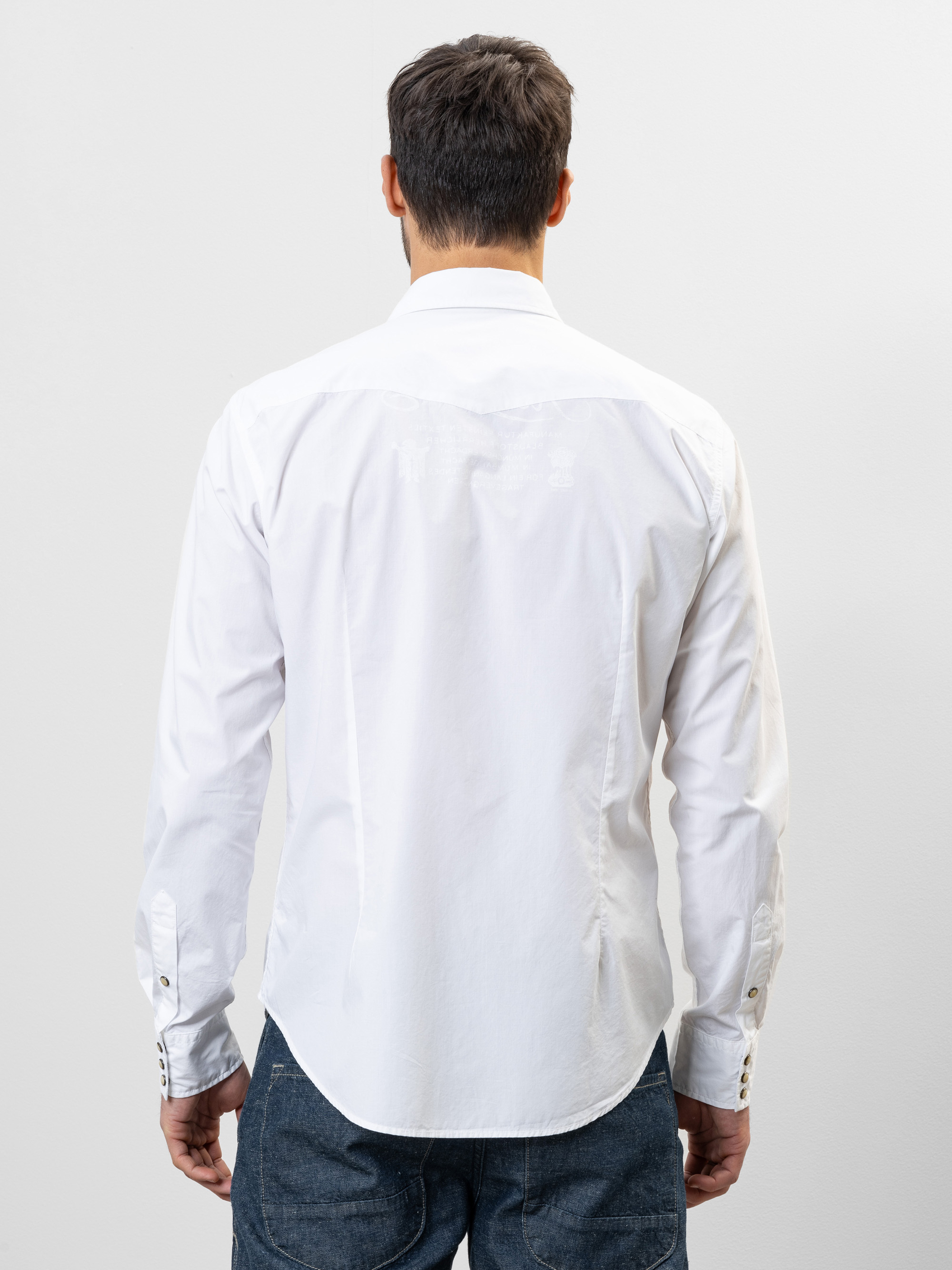 Ein Mann steht mit dem Rücken zur Kamera, trägt ein langärmeliges weißes Button-up-Hemd und blaue Jeans vor einem schlichten hellen Hintergrund.