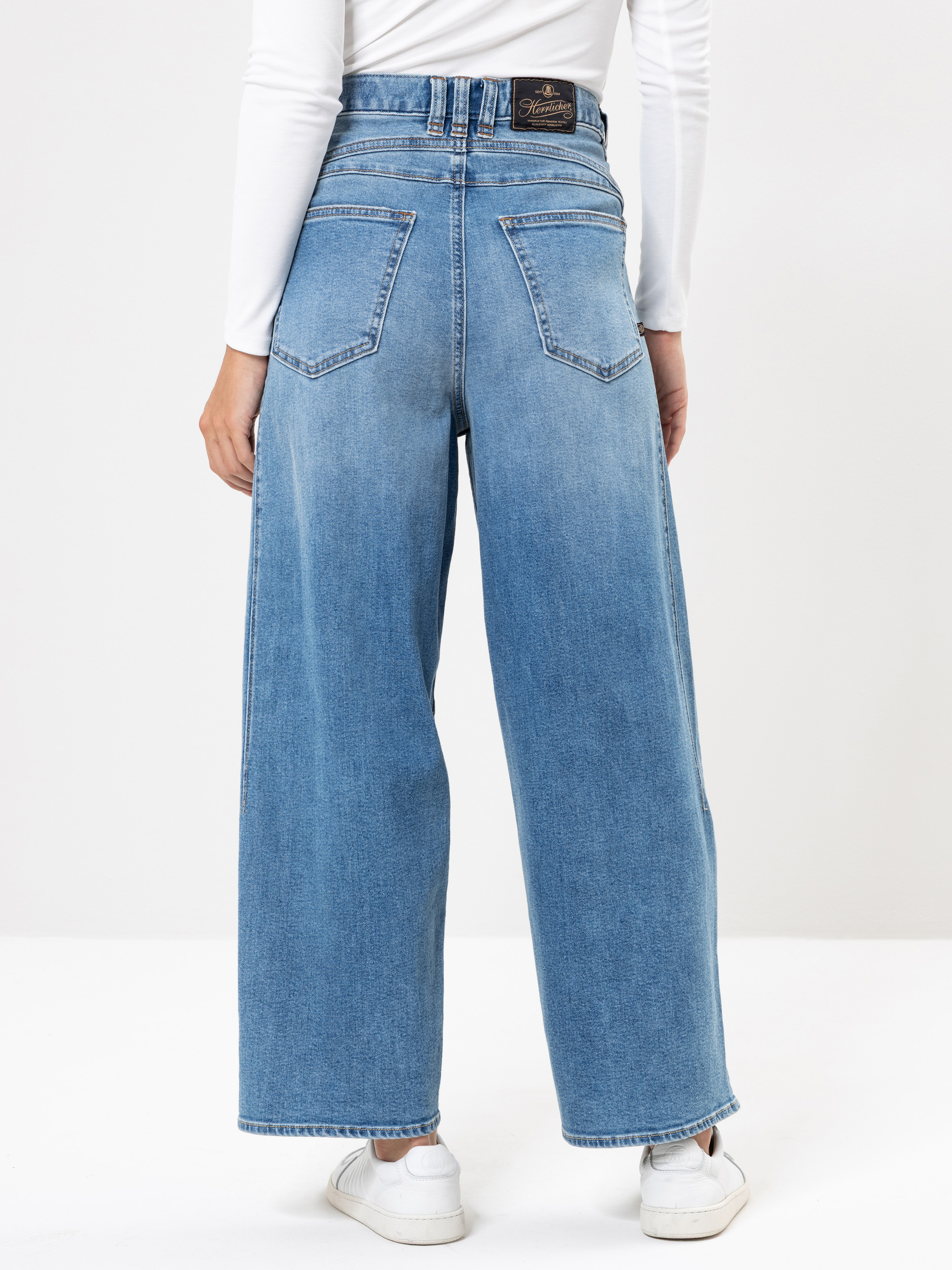 Eine Person, die eine blaue Jeans mit hoher Taille und weitem Bein, ein weißes langärmeliges Hemd und weiße Turnschuhe trägt, wird von hinten vor einem einfarbigen Hintergrund gezeigt.