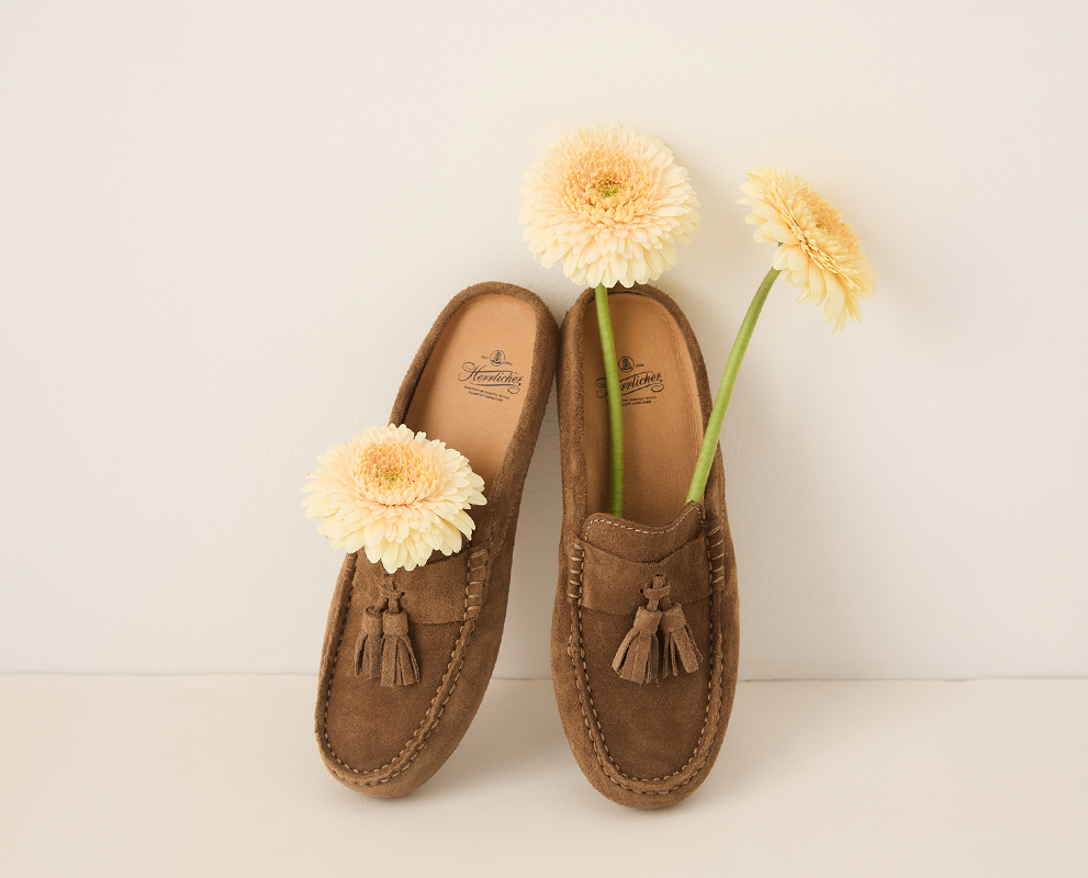 In einem Paar brauner Wildlederschuhe mit Quasten sind hellgelbe Blumen platziert, die aufrecht vor einem schlichten cremefarbenen Hintergrund stehen.