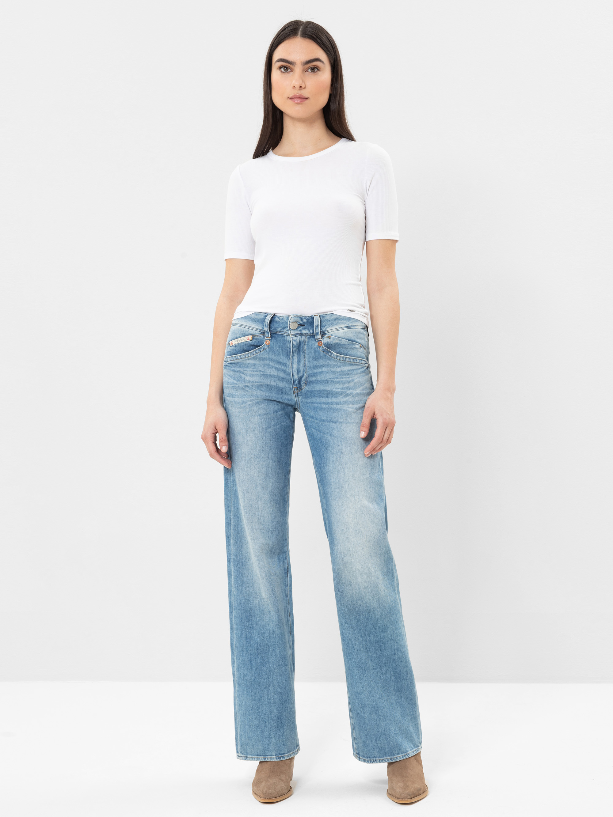 Eine Frau steht vor einem schlichten weißen Hintergrund und trägt ein weißes, kurzärmeliges Oberteil, hellblaue Jeans mit hoher Taille und weiten Beinen sowie beigefarbene Schuhe mit Absätzen. Sie schaut mit neutralem Blick direkt in die Kamera.
