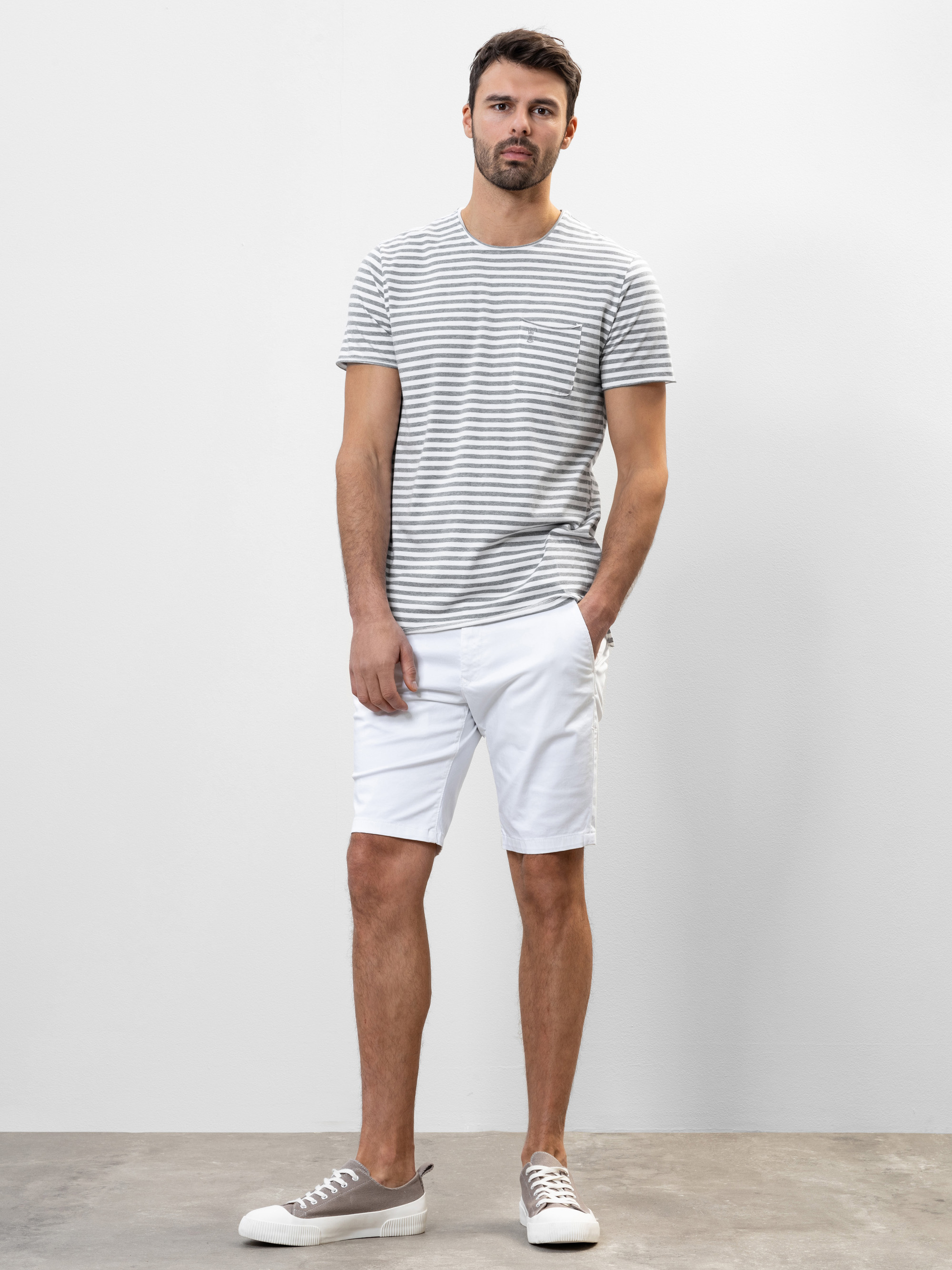Ein Mann steht vor einer schlichten weißen Wand und trägt ein grau-weiß gestreiftes T-Shirt, weiße Shorts und graue Turnschuhe. Eine Hand steckt in seiner Tasche und er hat einen neutralen Gesichtsausdruck.