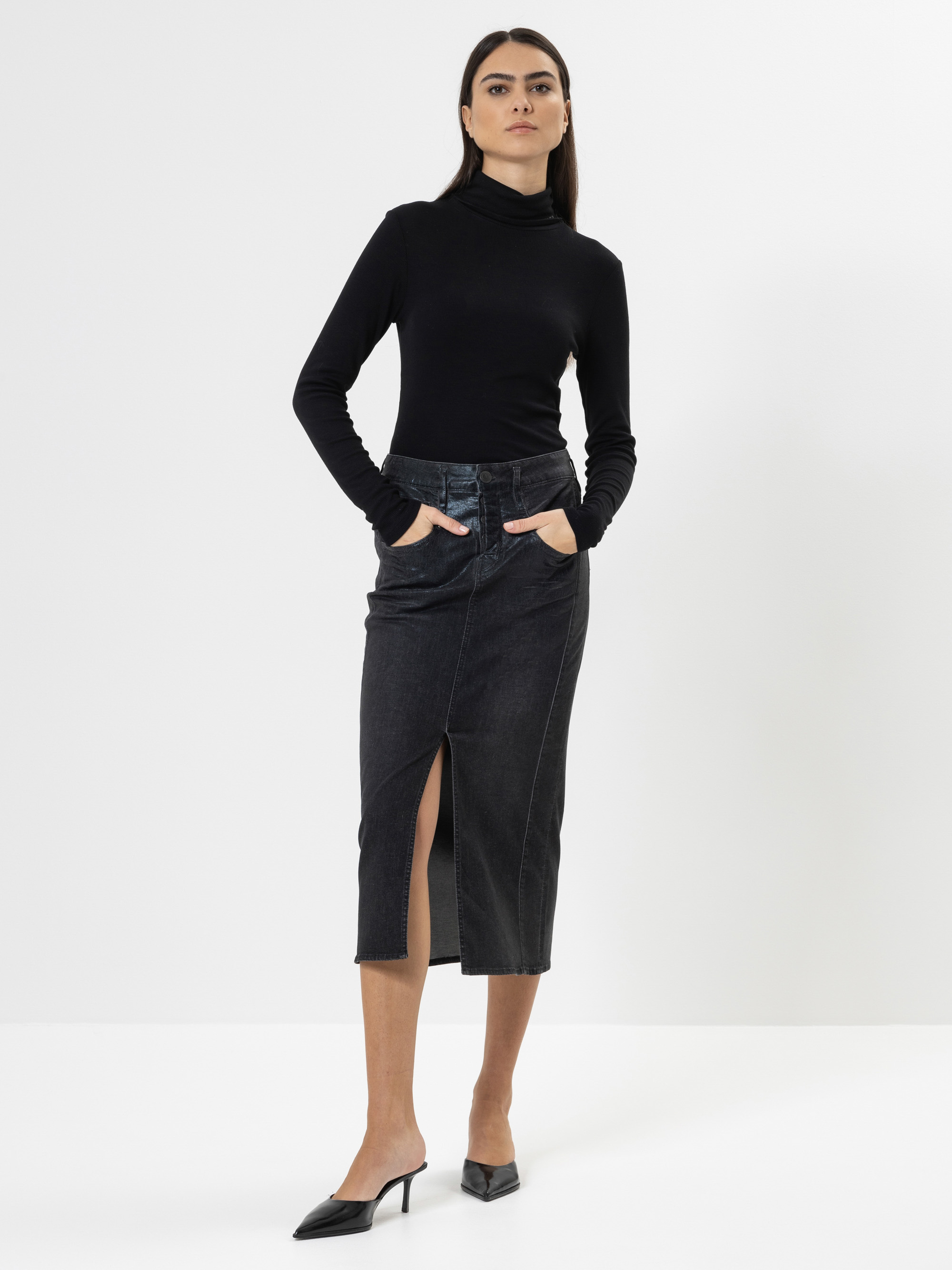 Eine Frau steht vor einem weißen Hintergrund und trägt einen schwarzen Rollkragenpullover, einen hochtaillierten dunklen Jeansrock mit einem Schlitz an der Vorderseite und schwarze Pantoletten mit Absatz. Sie hat die Hände in ihren Rocktaschen und eine