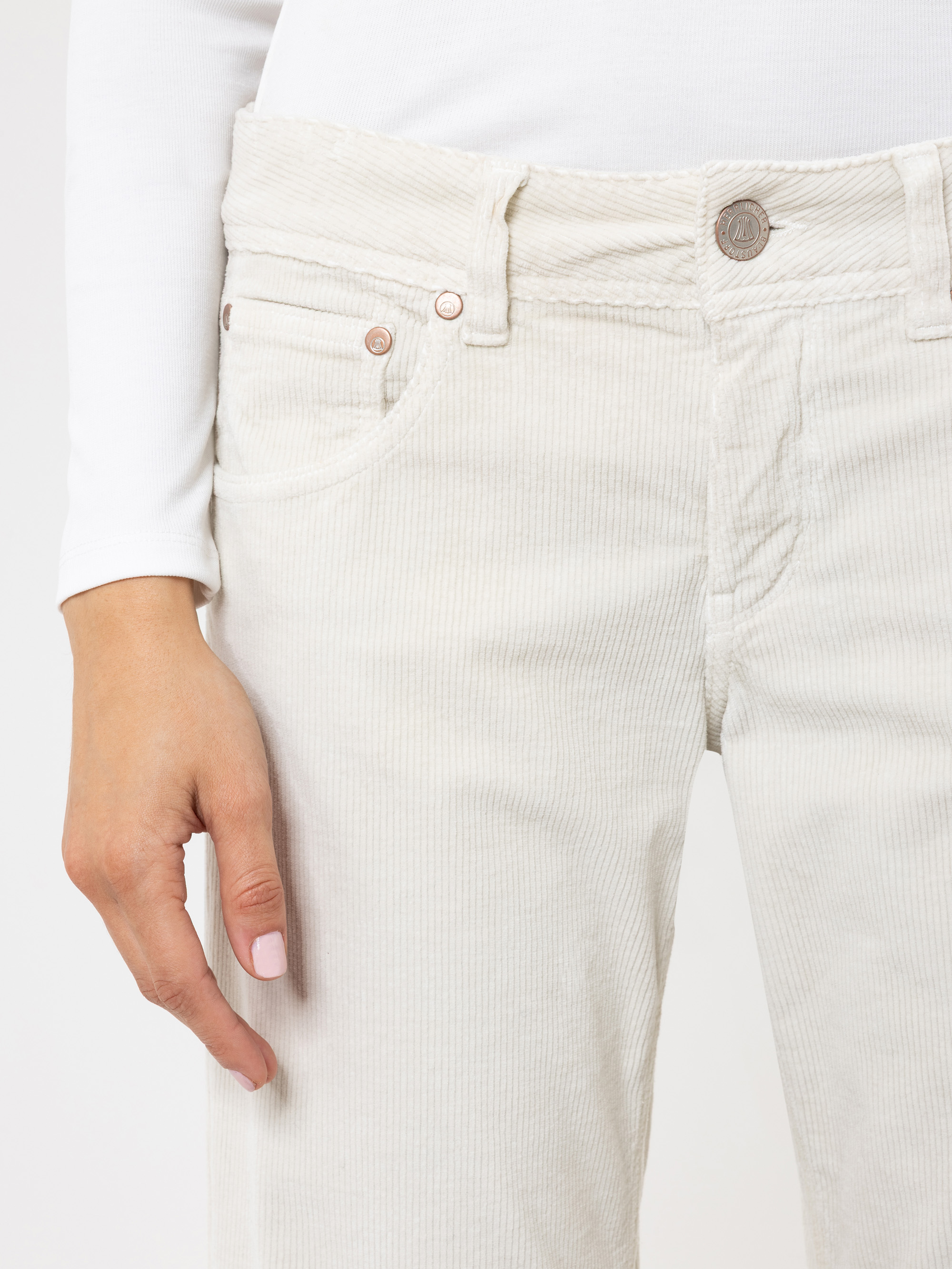 Eine Person, die eine cremefarbene Cordhose und ein weißes langärmeliges Oberteil trägt und ihre linke Hand an der Seite ruhen lässt. Der Fokus liegt auf der Hose und der Hand der Person.