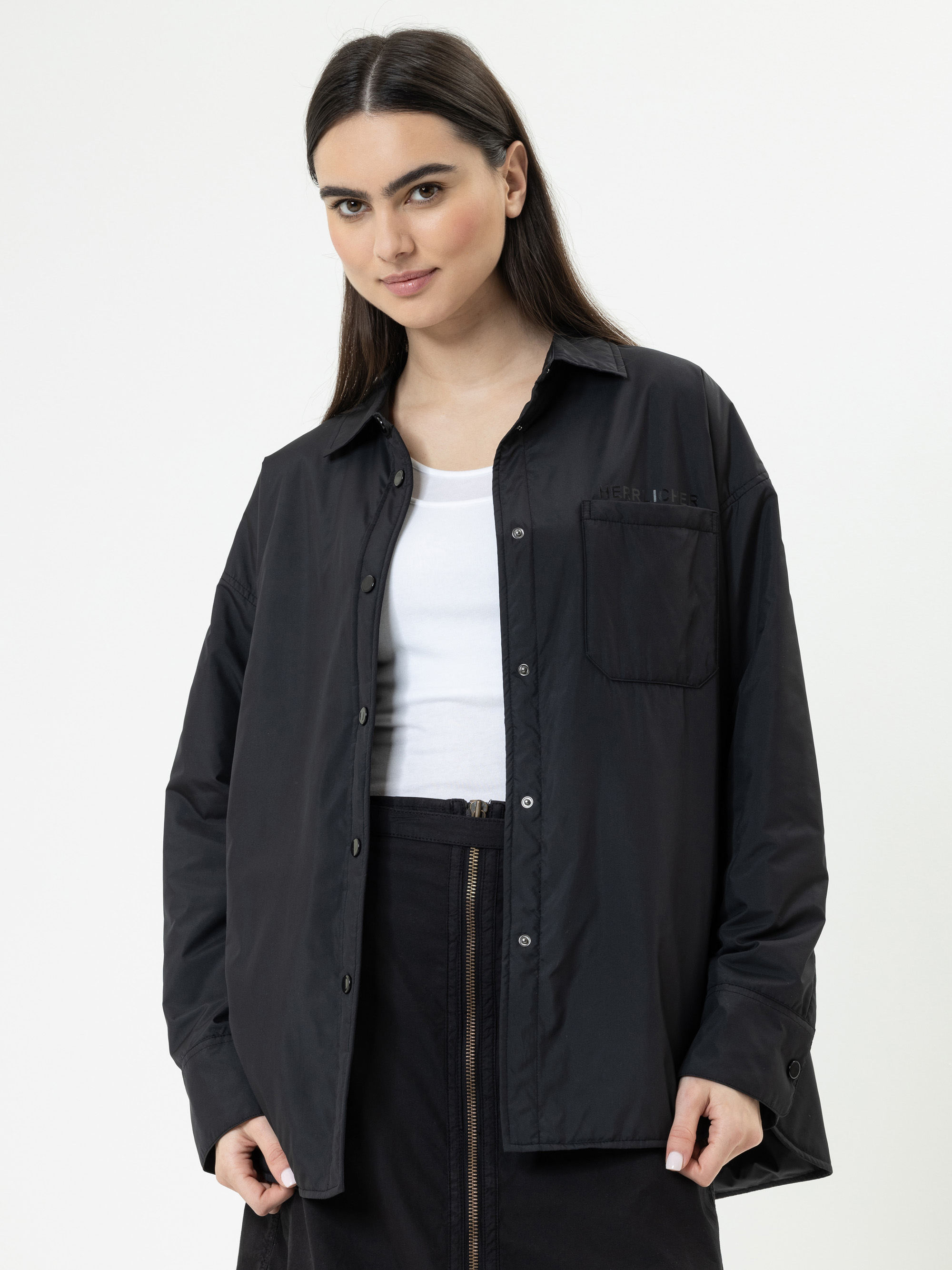 Eine Frau mit langen dunklen Haaren, die ein schwarzes Button-up-Hemd über einem weißen Oberteil und eine schwarze Hose trägt, steht vor einem schlichten hellen Hintergrund und schaut mit einem leichten Lächeln in die Kamera.