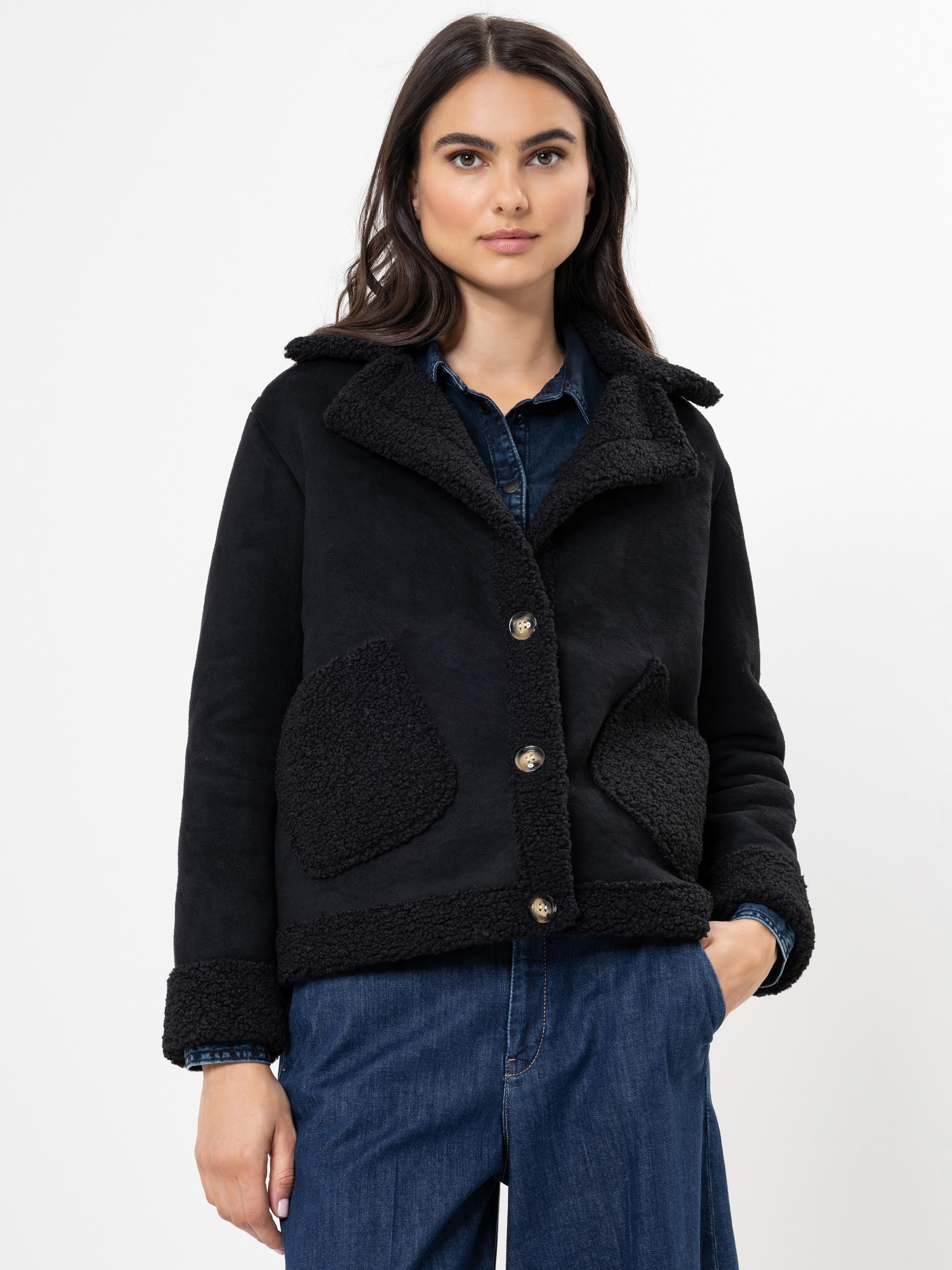 Eine Frau mit langen dunklen Haaren trägt eine schwarze geknöpfte Shearling-Jacke mit großen strukturierten Taschen und Manschetten, gepaart mit einem blauen Jeanshemd und blauen Jeans, vor einem schlichten weißen Hintergrund.
