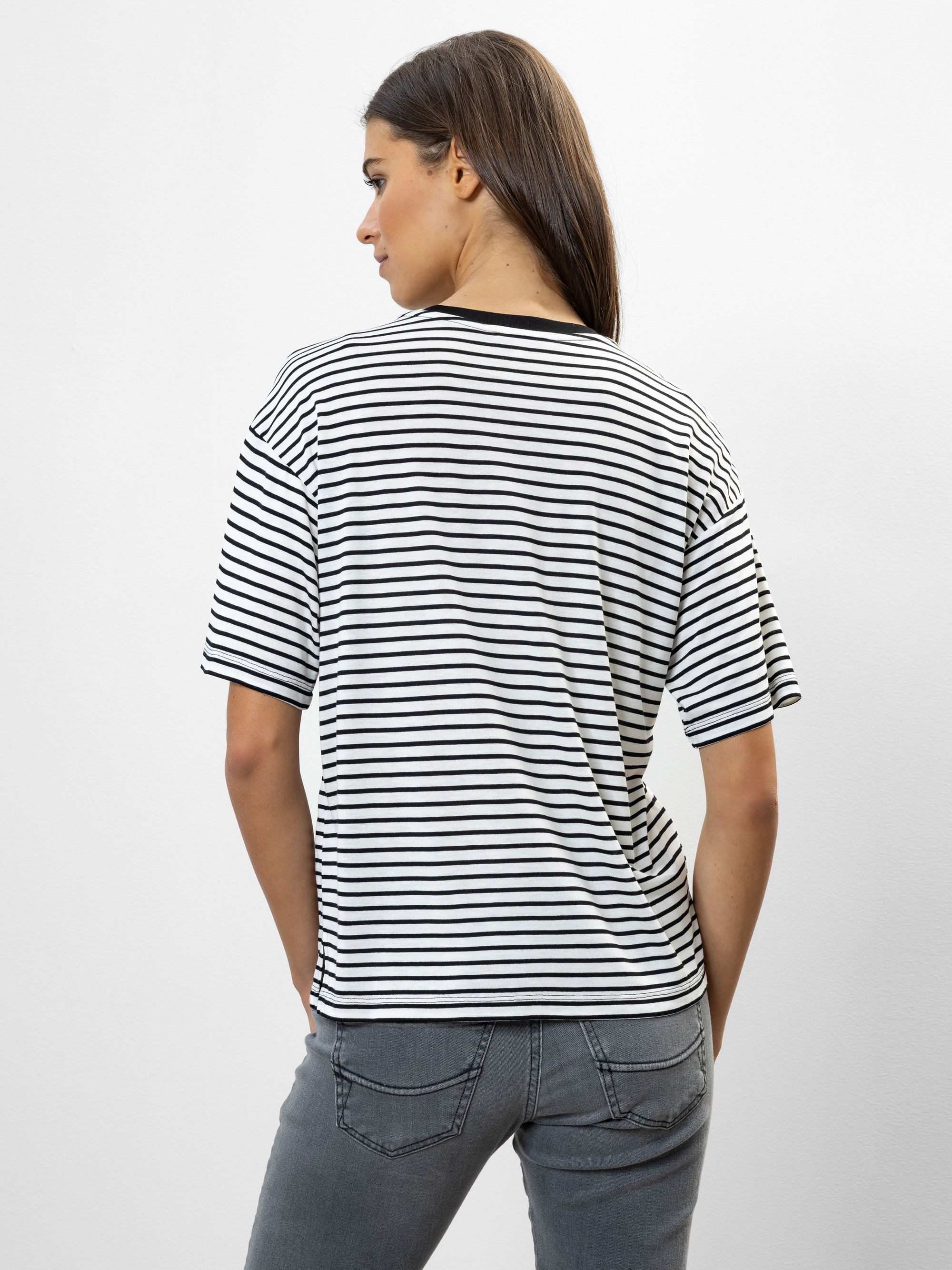 Eine Frau steht mit dem Gesicht zur Wand und trägt ein schwarz-weiß gestreiftes kurzärmeliges Hemd und graue Jeans vor einem schlichten weißen Hintergrund.