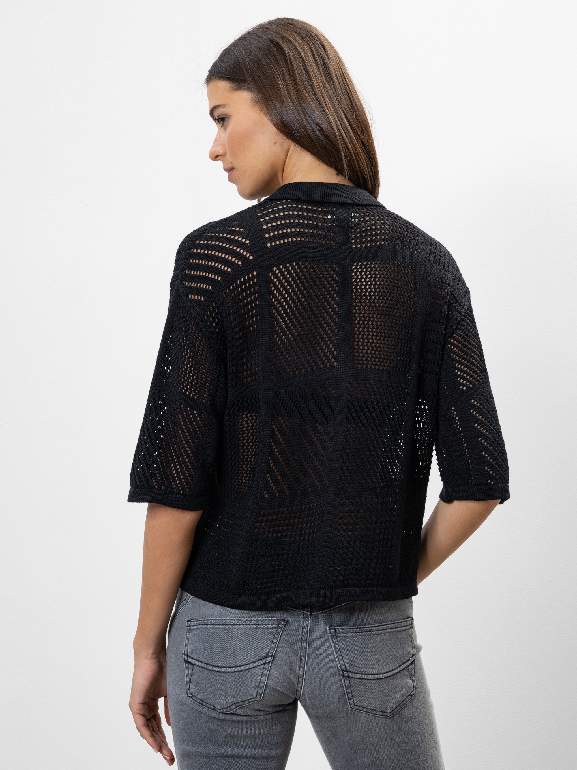 Eine Frau mit langen braunen Haaren, die ein schwarzes, kurzärmeliges, halbdurchsichtiges Strickoberteil und graue Jeans trägt, steht mit dem Rücken zur Kamera vor einem schlichten weißen Hintergrund.