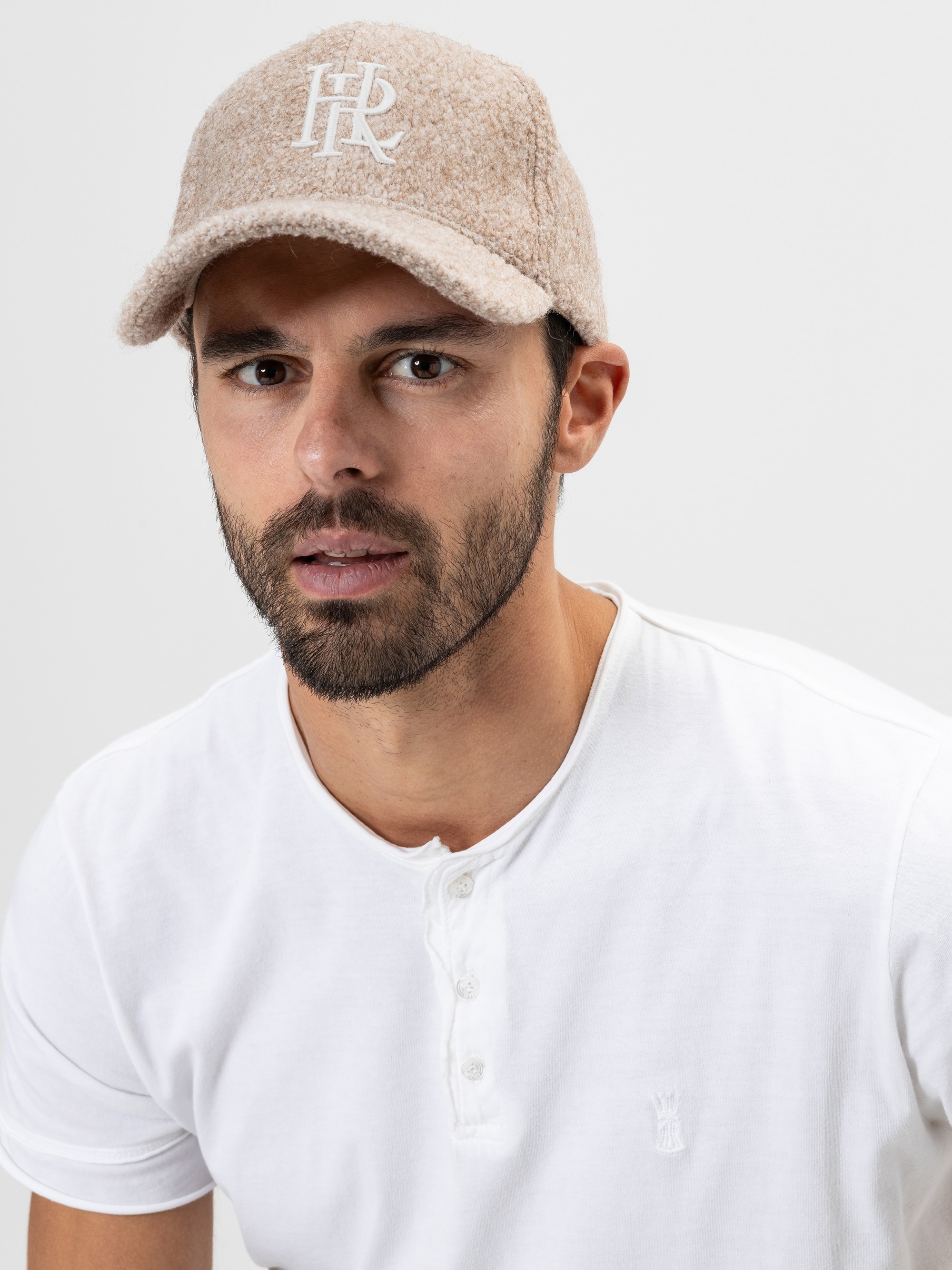 Ein Mann mit kurzem Bart und Schnurrbart trägt eine hellbeige, strukturierte Mütze mit weißen aufgestickten Buchstaben und ein weißes, kurzärmeliges Hemd, das sich vor einem schlichten, hellen Hintergrund befindet.