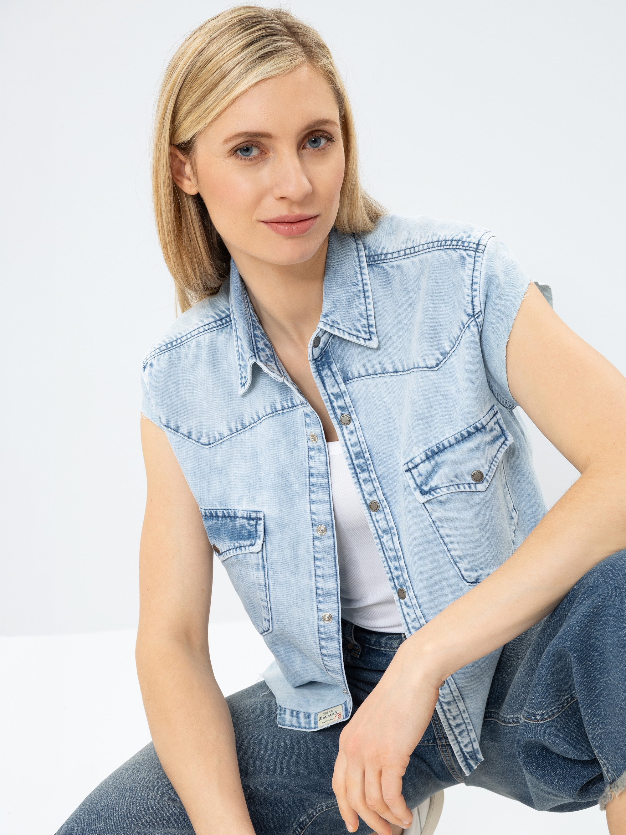 Eine Frau mit glattem blondem Haar, die ein ärmelloses helles Jeanshemd über einem weißen Oberteil und blaue Jeans trägt, sitzt vor einem schlichten Hintergrund und lächelt sanft in die Kamera.