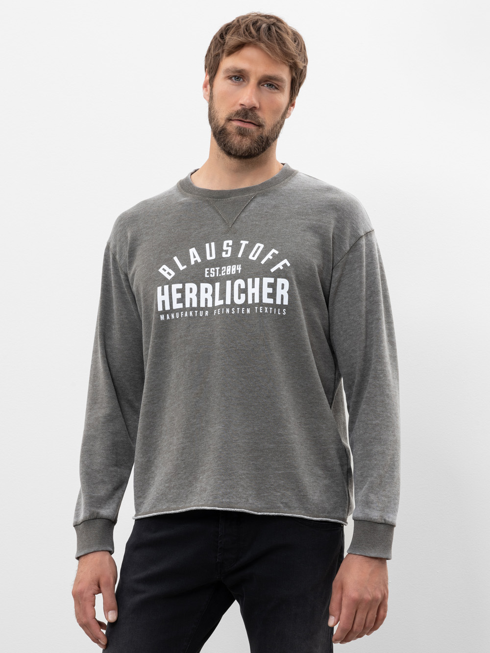 Ein Mann mit kurzen braunen Haaren und einem Bart trägt ein locker sitzendes, graues, langärmeliges Sweatshirt mit der weißen Aufschrift "BLAUSTOFF HERRLICHER" und schwarze Jeans vor einem schlichten, hellen Hintergrund.
