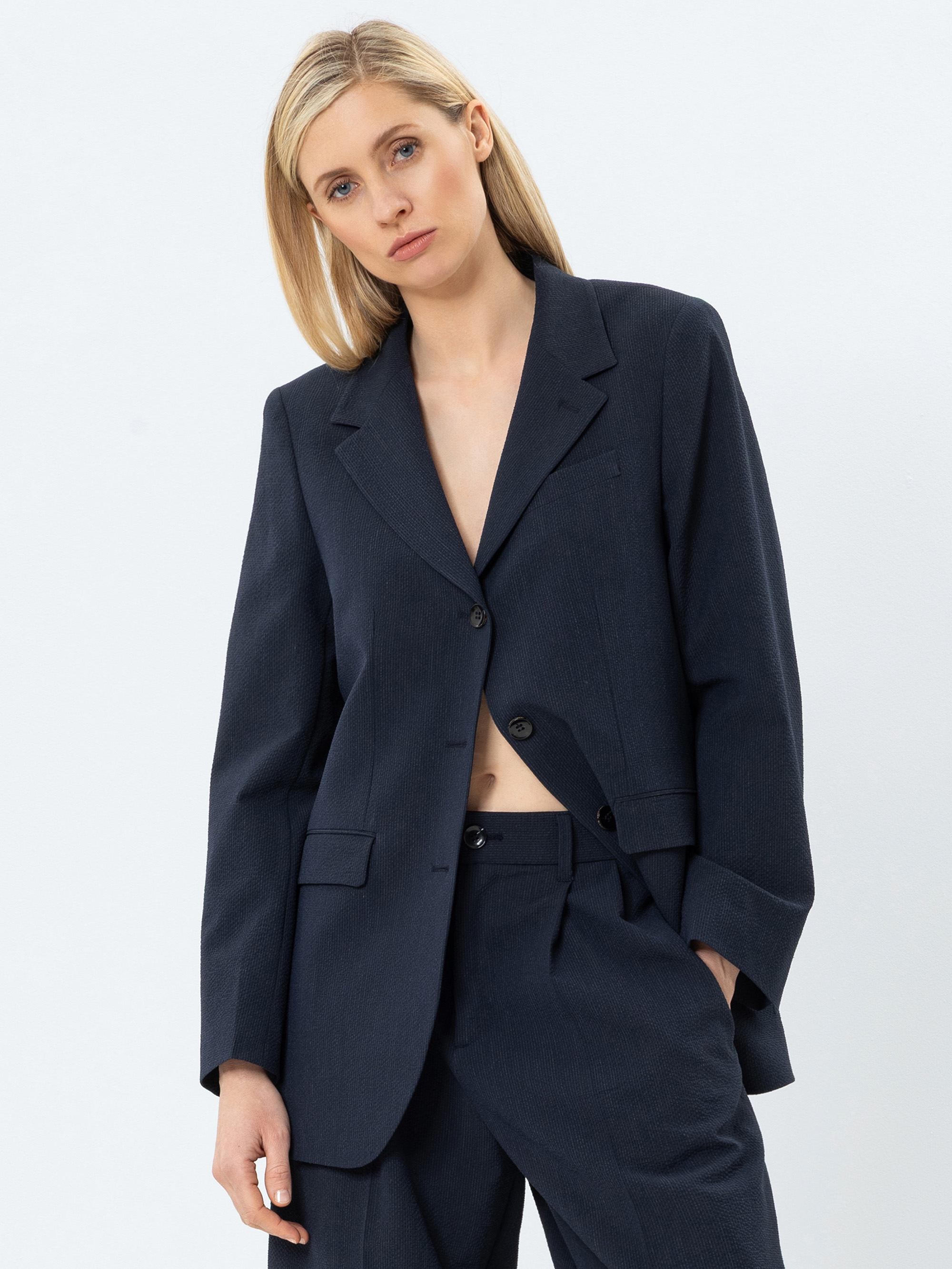 Eine Frau mit glattem blondem Haar, die eine dunkle marineblaue Anzugsjacke und eine dazu passende Hose trägt, steht vor einem schlichten weißen Hintergrund und schaut mit neutralem Blick in die Kamera.