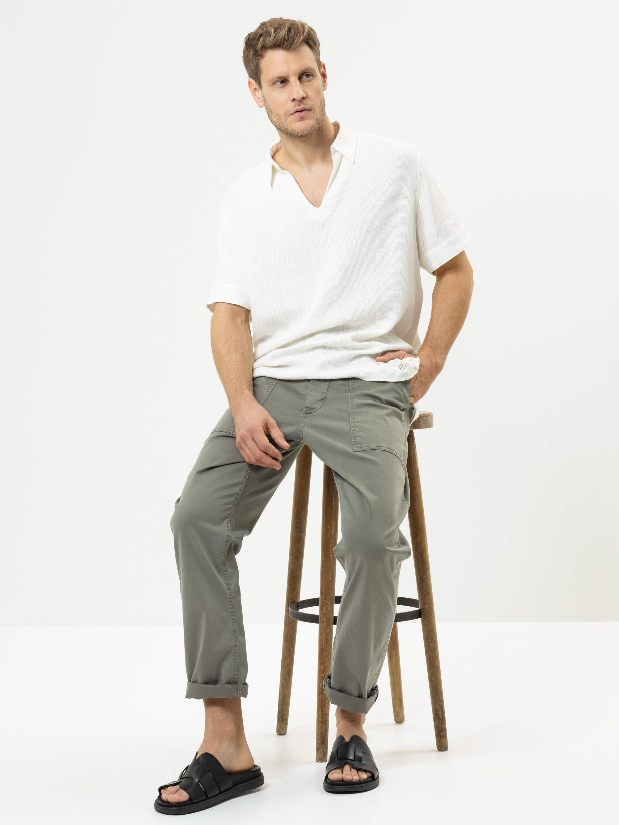 Ein Mann mit kurzen Haaren sitzt auf einem Holzschemel vor einem schlichten weißen Hintergrund. Er trägt ein weißes kurzärmeliges Hemd, eine hellolivgrüne Hose und schwarze Sandalen. Er nimmt eine entspannte Haltung ein und hat eine Hand in seiner Ta