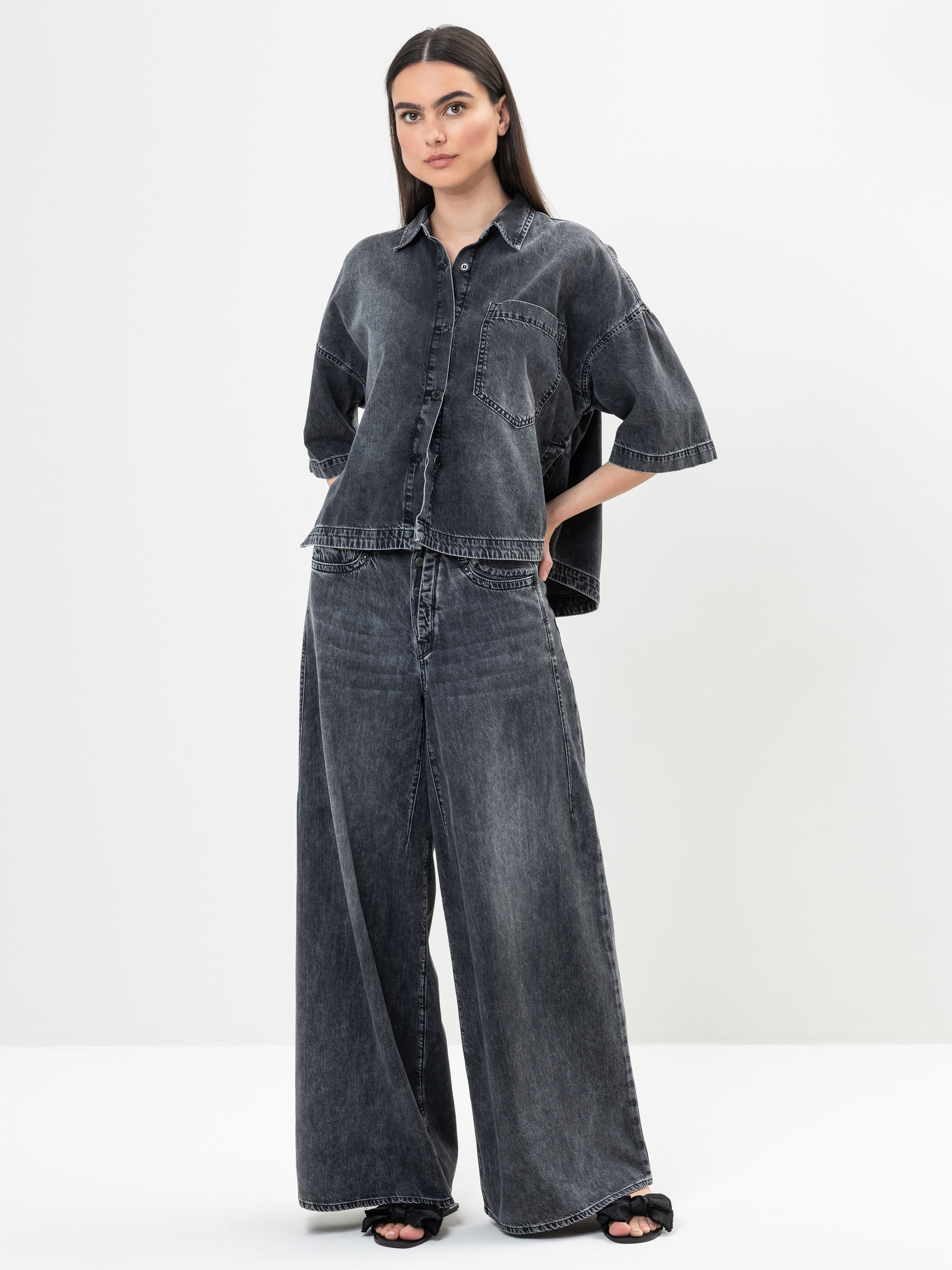 Eine Frau steht vor einem schlichten weißen Hintergrund und trägt ein locker sitzendes, dunkel gewaschenes Jeanshemd mit kurzen Ärmeln und eine dazu passende Jeanshose mit weitem Bein. Sie hat langes dunkles Haar und trägt schwarze offene Sandalen.