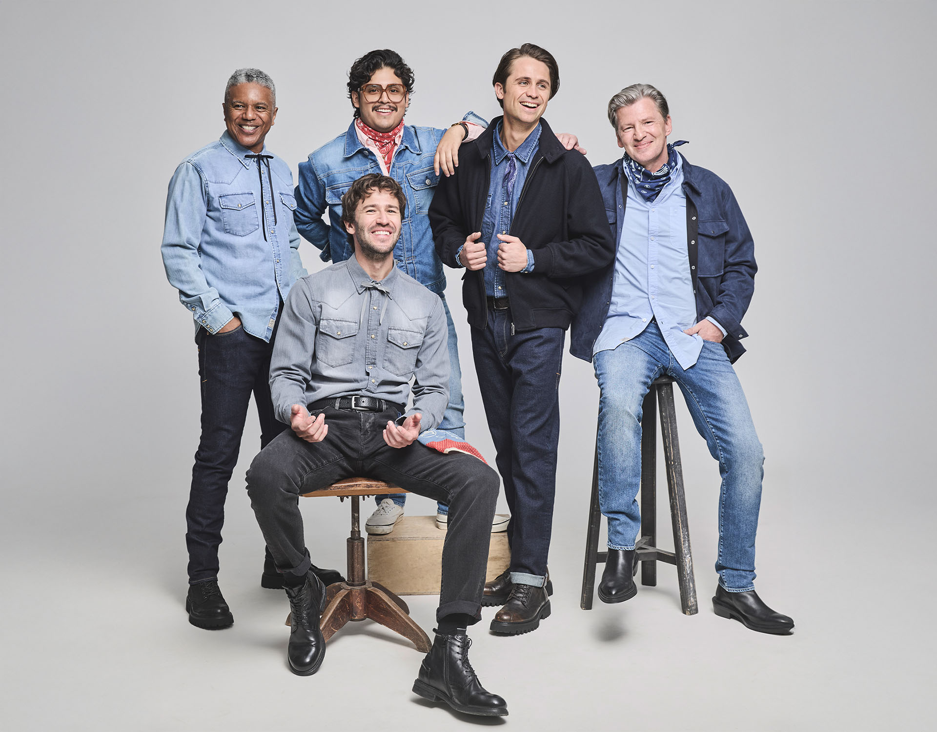 Fünf lächelnde Männer in Jeans und Freizeitkleidung posieren zusammen in einem Studio. Vier stehen, einer sitzt auf einem Stuhl. Sie tragen blaue Jeans, Hemden, Jacken, Kopftücher und Stiefel vor einem schlichten hellen Hintergrund.