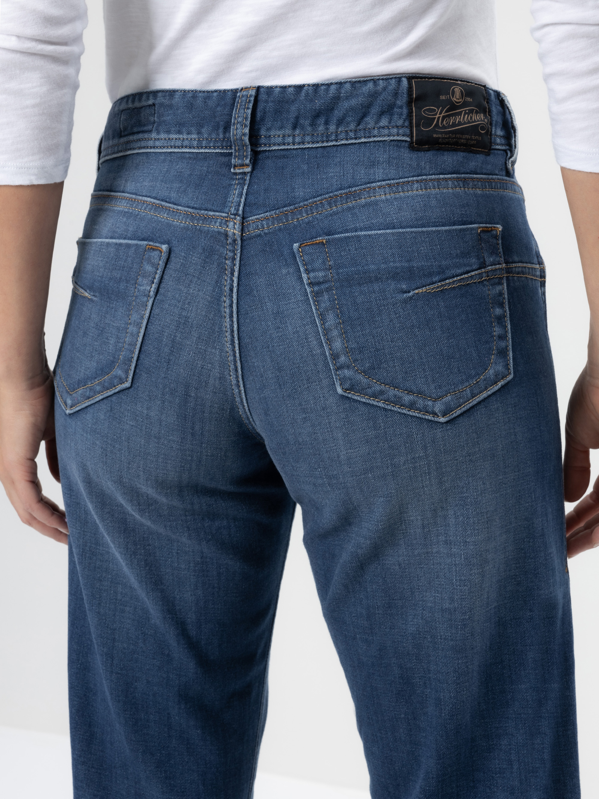 Eine Person trägt eine mittelblaue Jeans und ein weißes langärmeliges Hemd, von hinten gesehen. Die Jeans hat zwei sichtbare Gesäßtaschen und einen schwarzen Markenaufnäher aus Leder über der rechten Tasche.