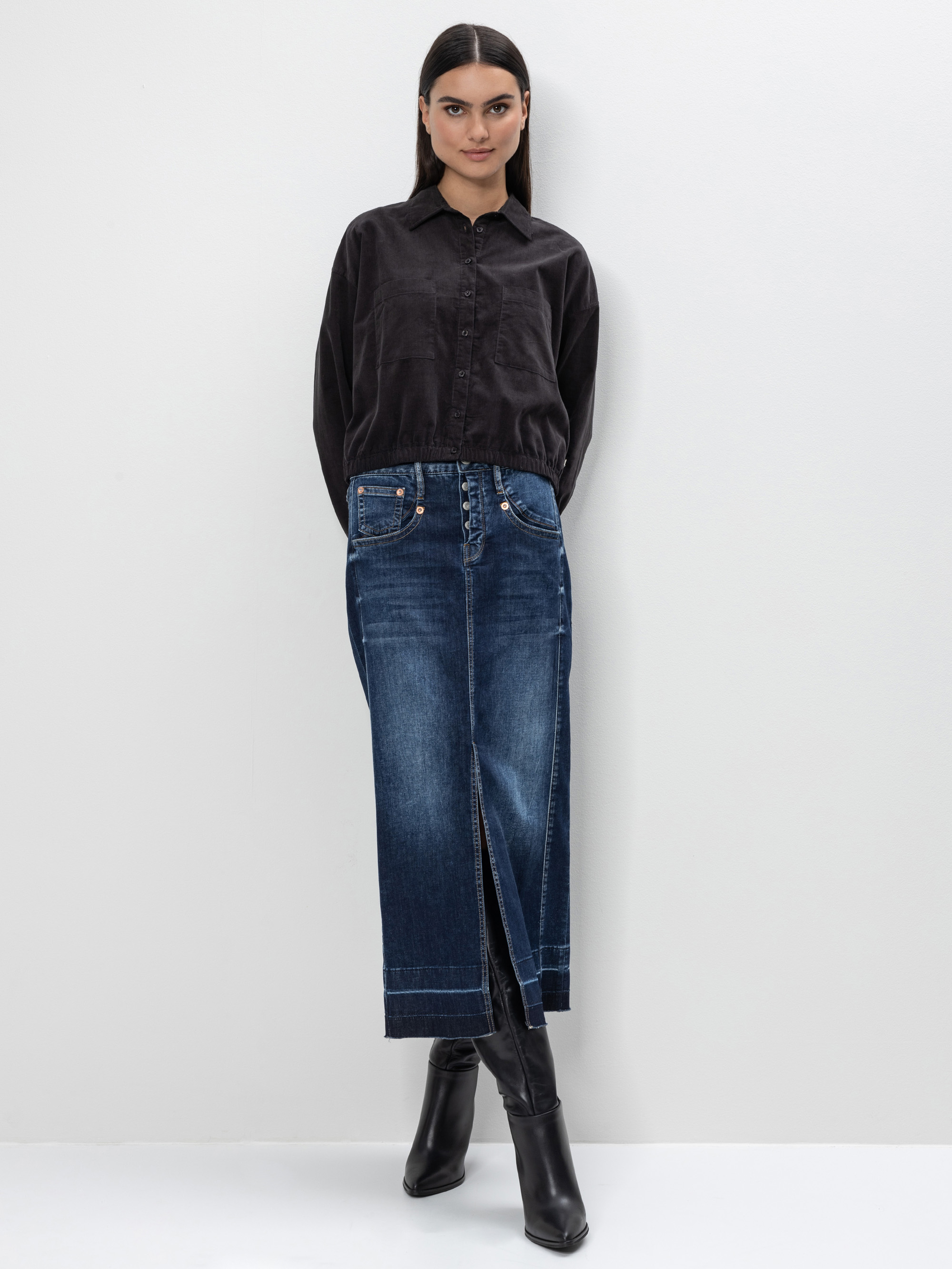 Eine Frau steht vor einer schlichten weißen Wand. Sie trägt ein schwarzes Button-up-Hemd, das in einen hochtaillierten, dunkelblauen Jeans-Maxirock mit einem vorderen Schlitz gesteckt wird. Sie hat glattes Haar und trägt schwarze hochhackige Stiefel.